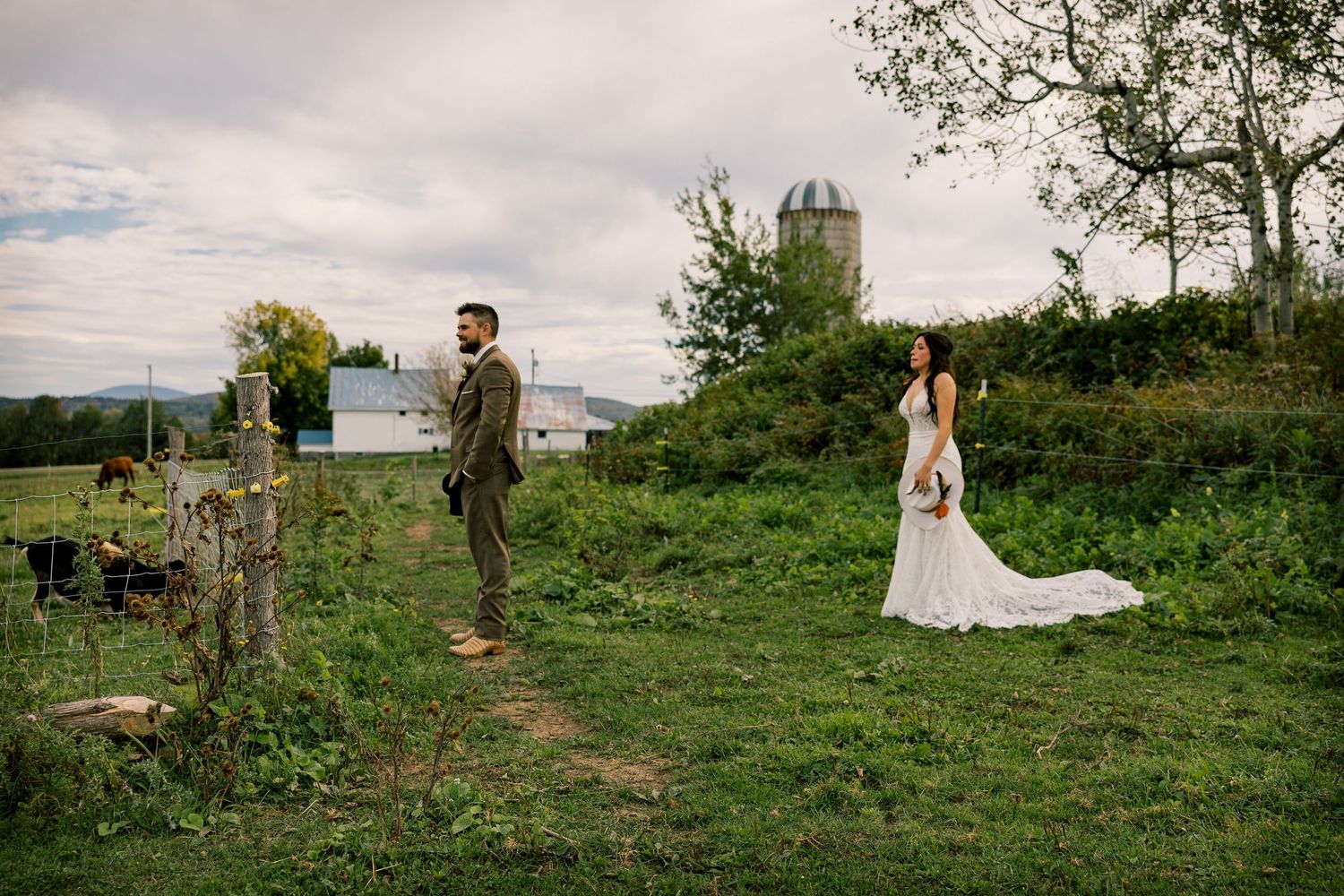 Bride in a long white gown and groom in a olive suit stand apart in a grassy field with a farmhouse, silo and cloudy sky behind them.