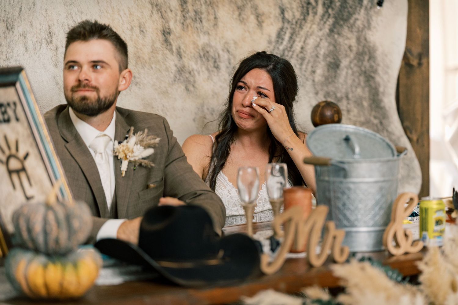 Emotional bride wipes a tear beside her smiling groom at a rustic wedding reception table with pumpkins, a cowboy hat and wooden letters.