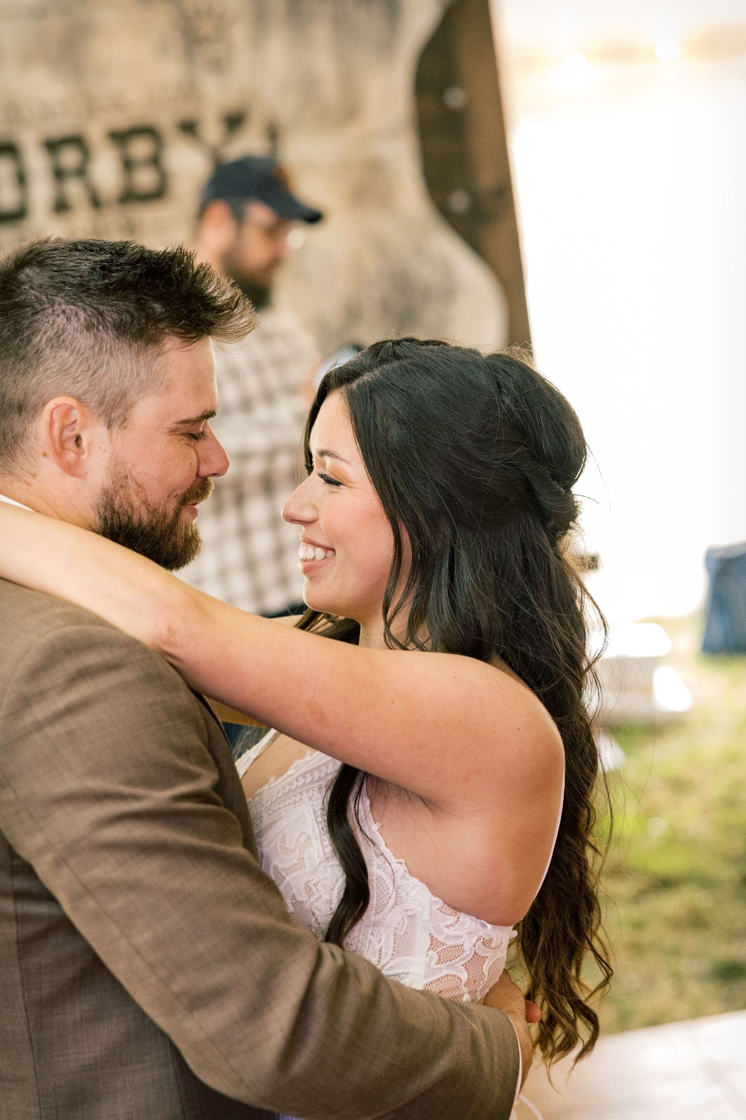 Smiling bride in a lace gown embraces a bearded groom in a brown jacket at an outdoor rustic wedding reception.