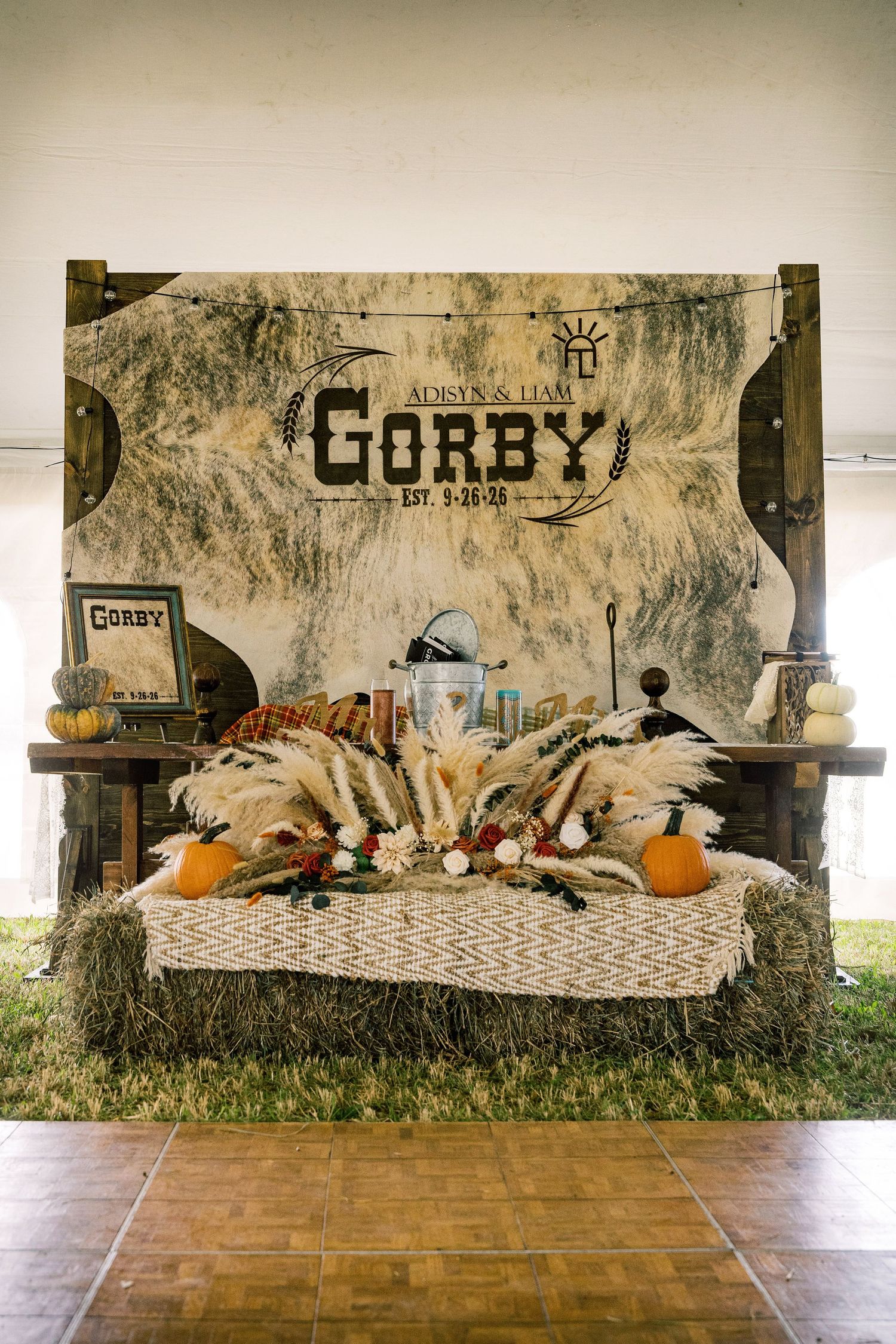 Rustic wedding sweetheart table on hay bales with pampas grass, pumpkins and a cowhide backdrop reading Gorby, set under a tent.