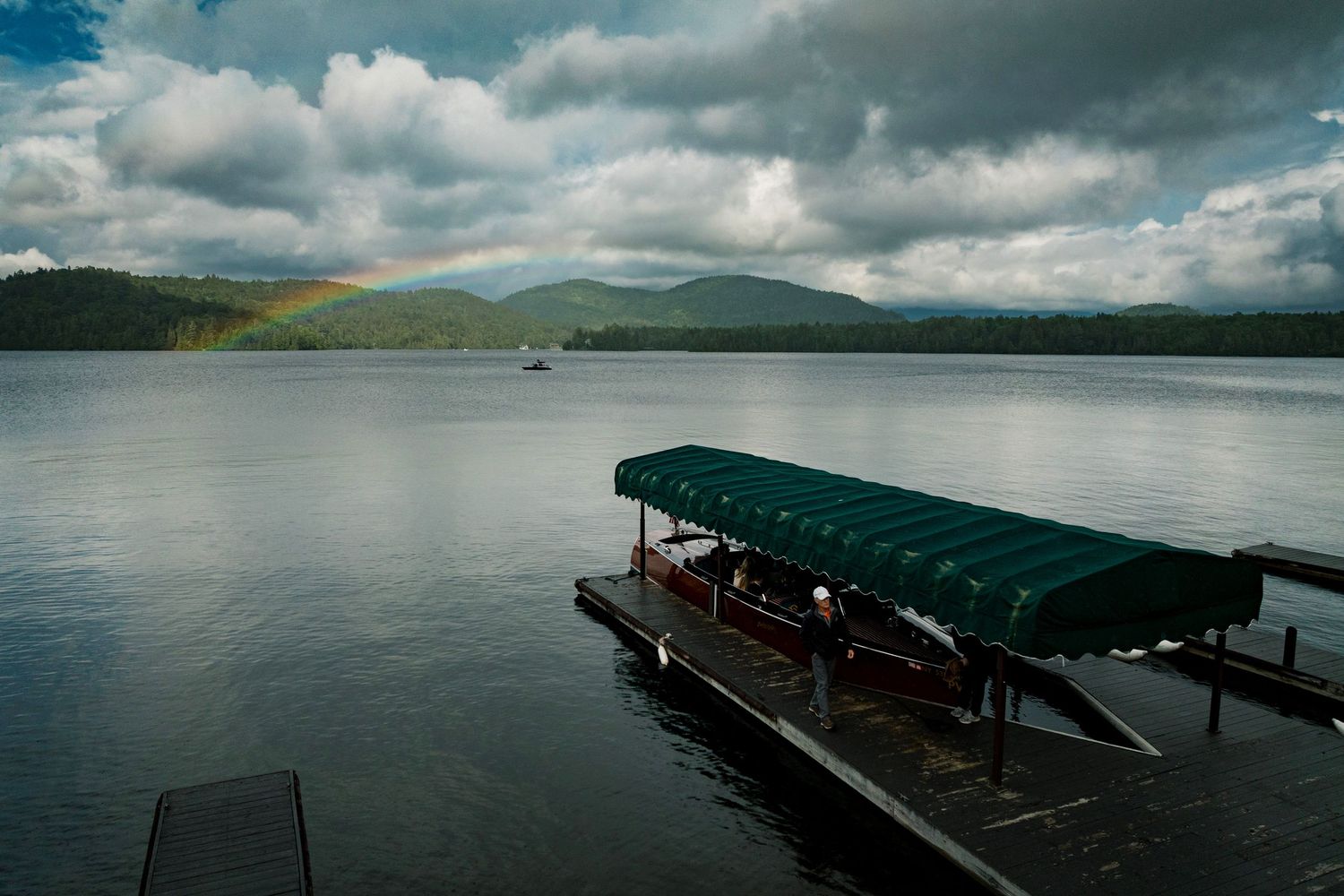 A covered boat dock on a calm lake with a rainbow over misty mountains under dramatic cloudy skies.