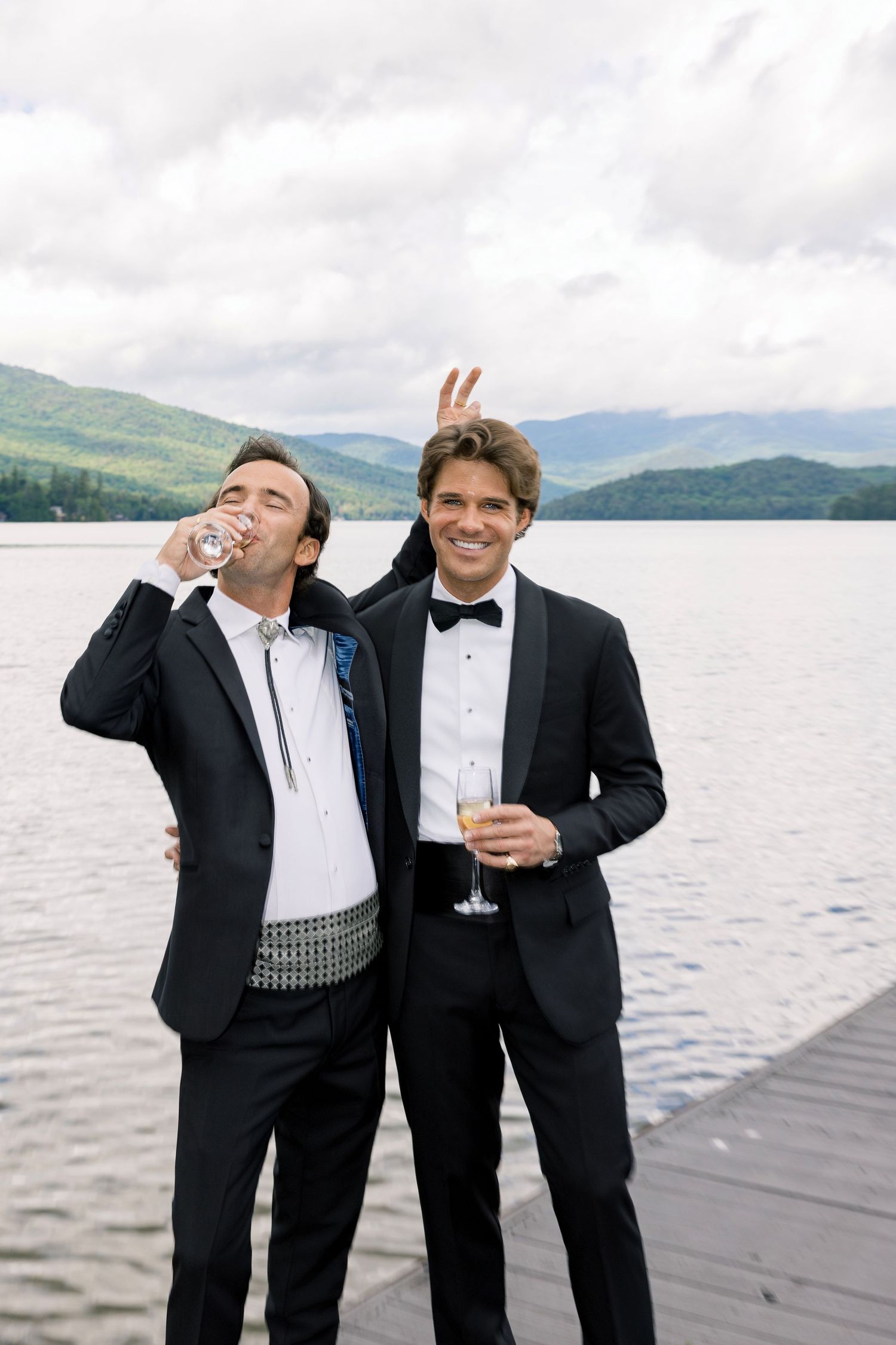 Two men in tuxedos celebrate on a lakeside dock, one drinking champagne while the other gives bunny ears.
