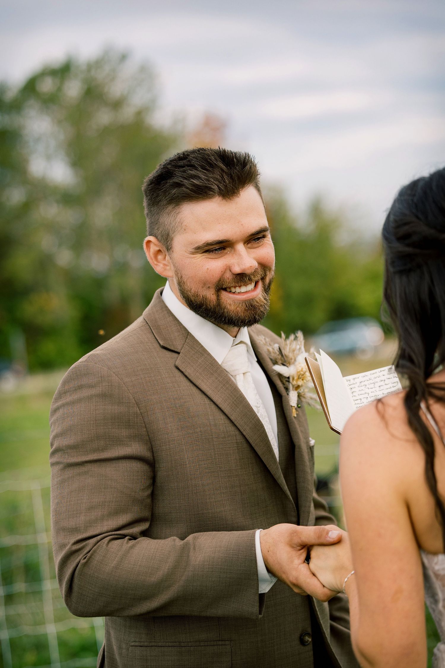 Smiling bearded groom in a brown suit with a floral boutonniere holds hands with his bride during an outdoor wedding ceremony.