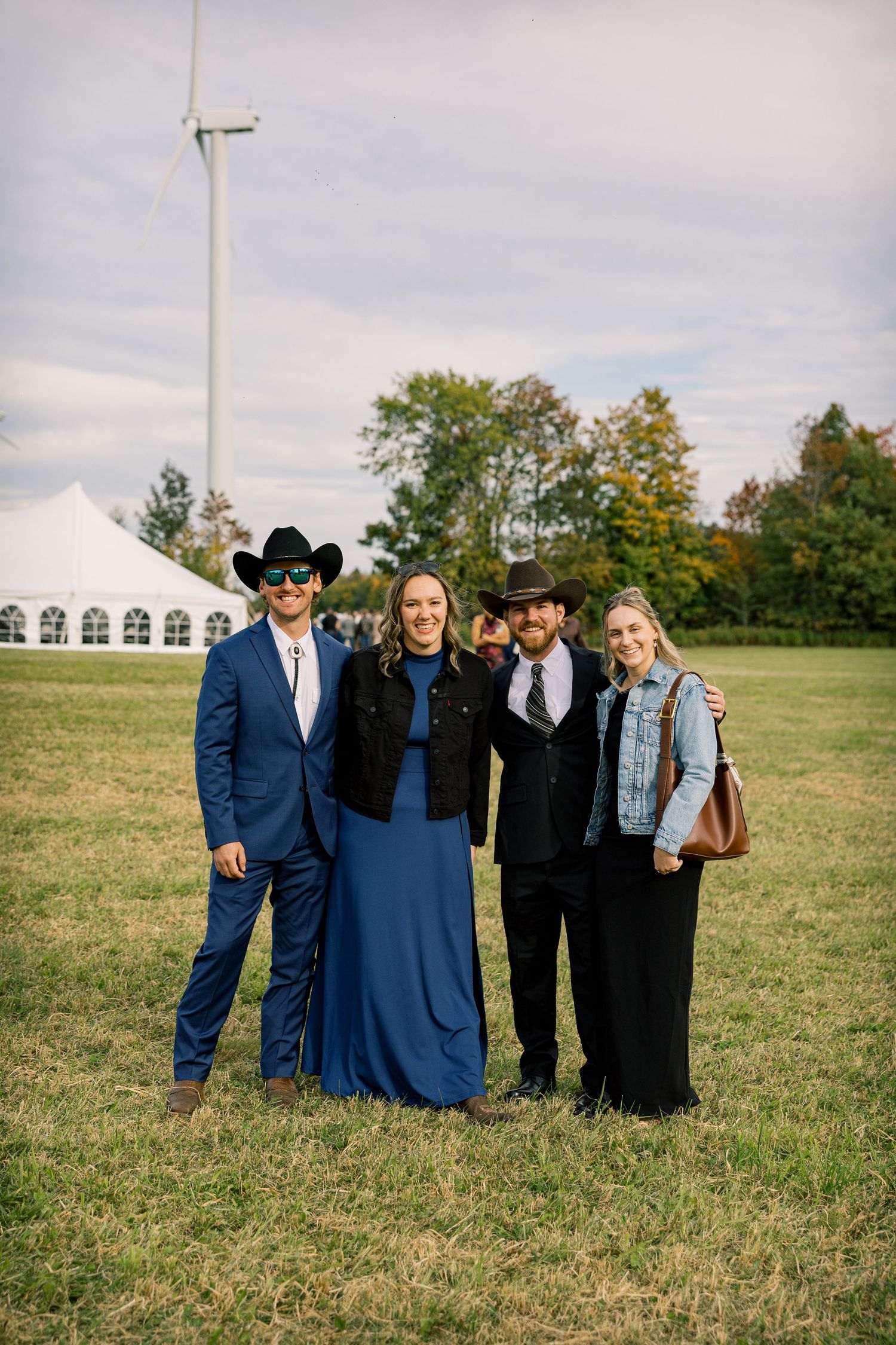 Four people in western wear, including cowboy hats and a blue dress, pose in a grassy field near a white tent and wind turbine.