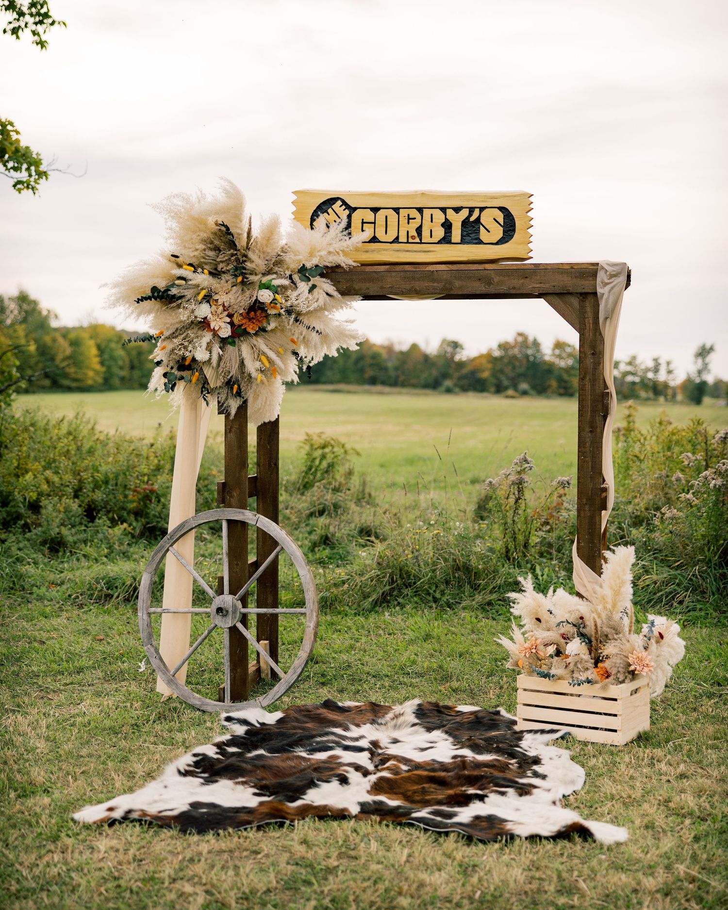 Rustic wooden Corby's sign arch with pampas grass florals, wagon wheel and cowhide rug in a grassy field, styled for a western-themed event.