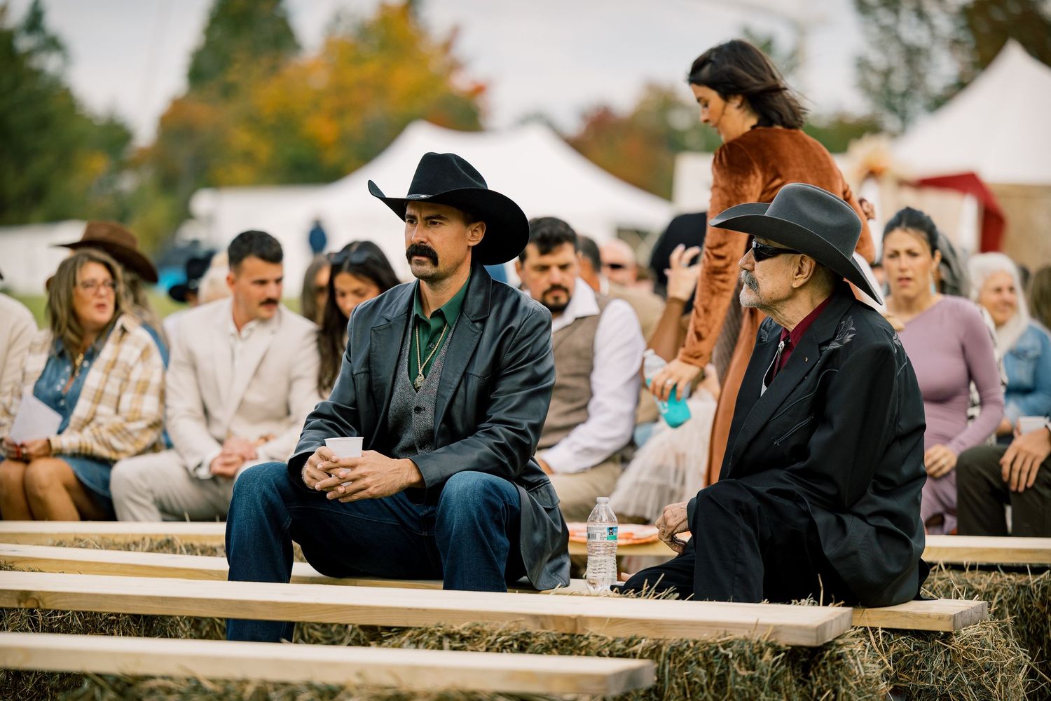 Two men in cowboy hats sit on hay bale seating at an outdoor western ceremony while guests gather behind them under white tents.