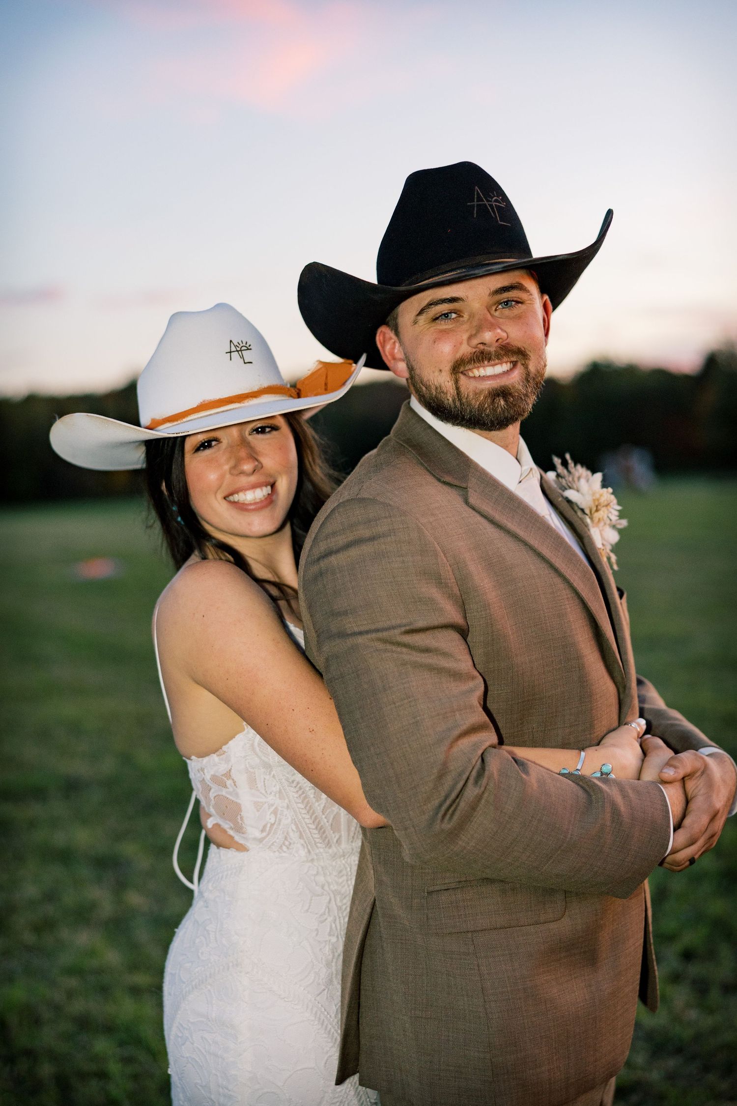 Smiling bride in a white lace dress and cowboy hat embraces a groom in a brown suit and black cowboy hat at sunset in a field.
