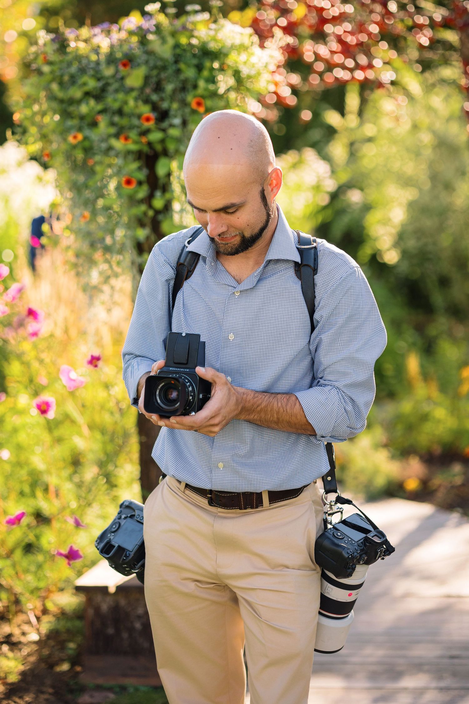 Bald male photographer reviewing camera in colorful garden, carrying multiple professional cameras and wearing blue shirt.