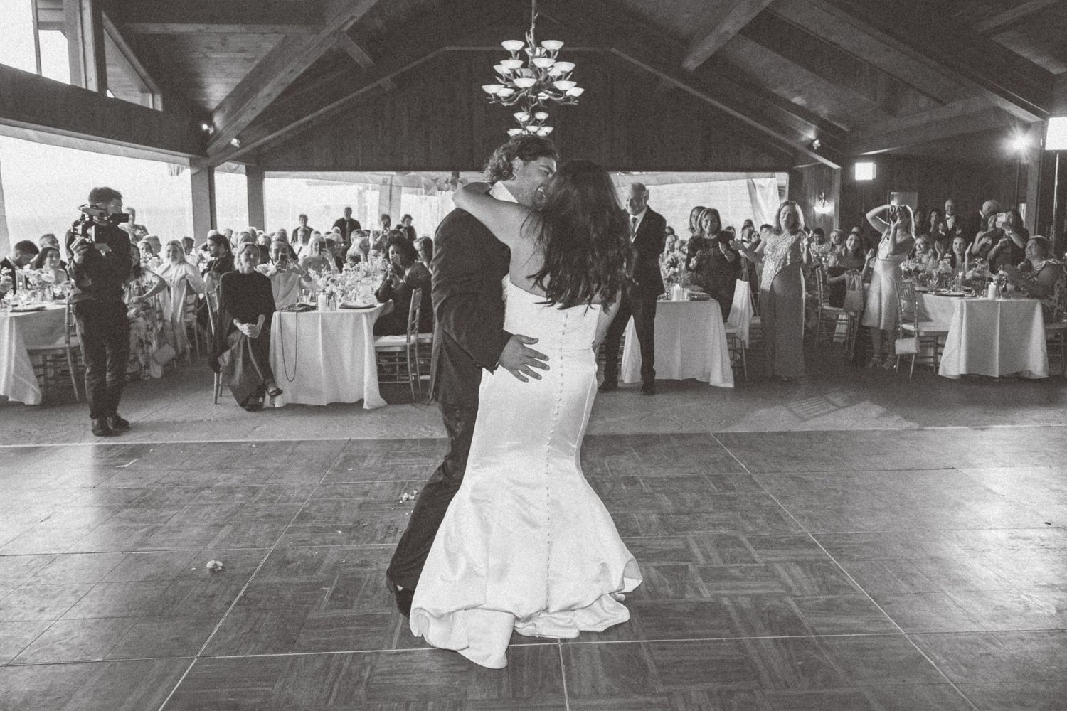 Black and white photo of bride and groom sharing first dance at wedding reception with guests watching in background.