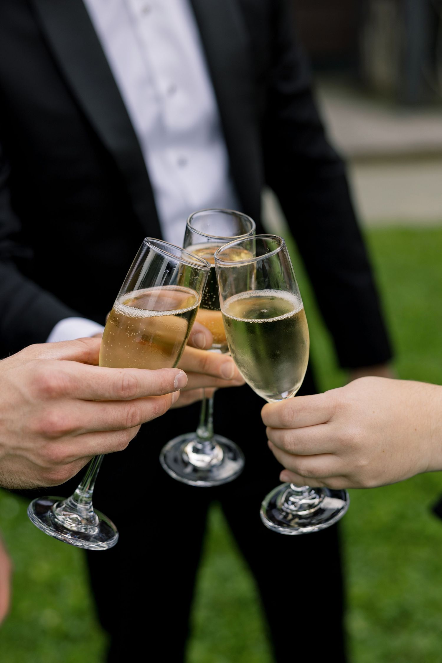 People clinking champagne flutes in a celebratory toast outdoors, with a man in a black tuxedo in the background.