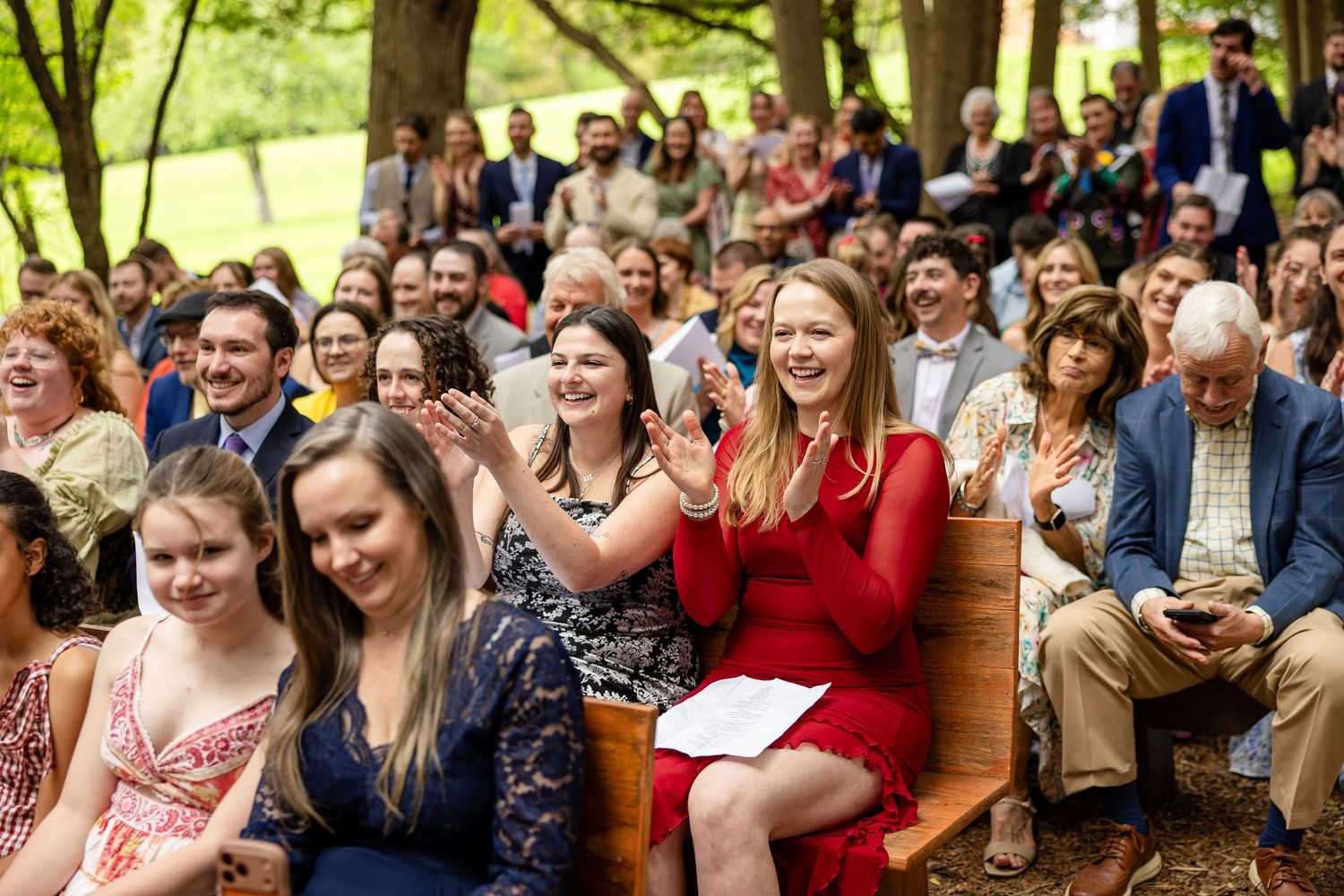 Guests clapping and smiling at an outdoor wedding ceremony seated on wooden benches surrounded by trees.