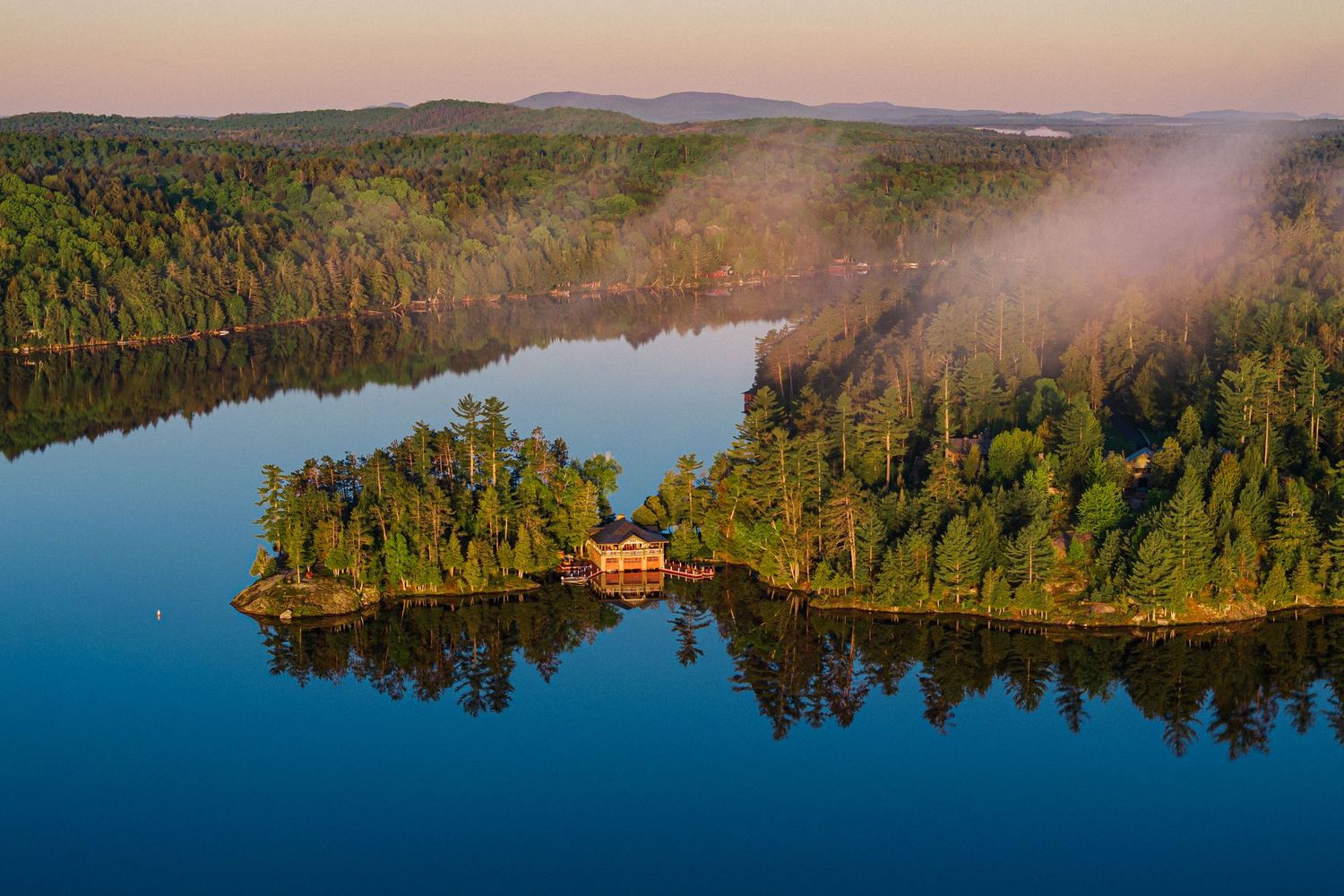 Aerial panoramic view of a red cabin nestled among pine trees beside a tranquil lake at sunset.