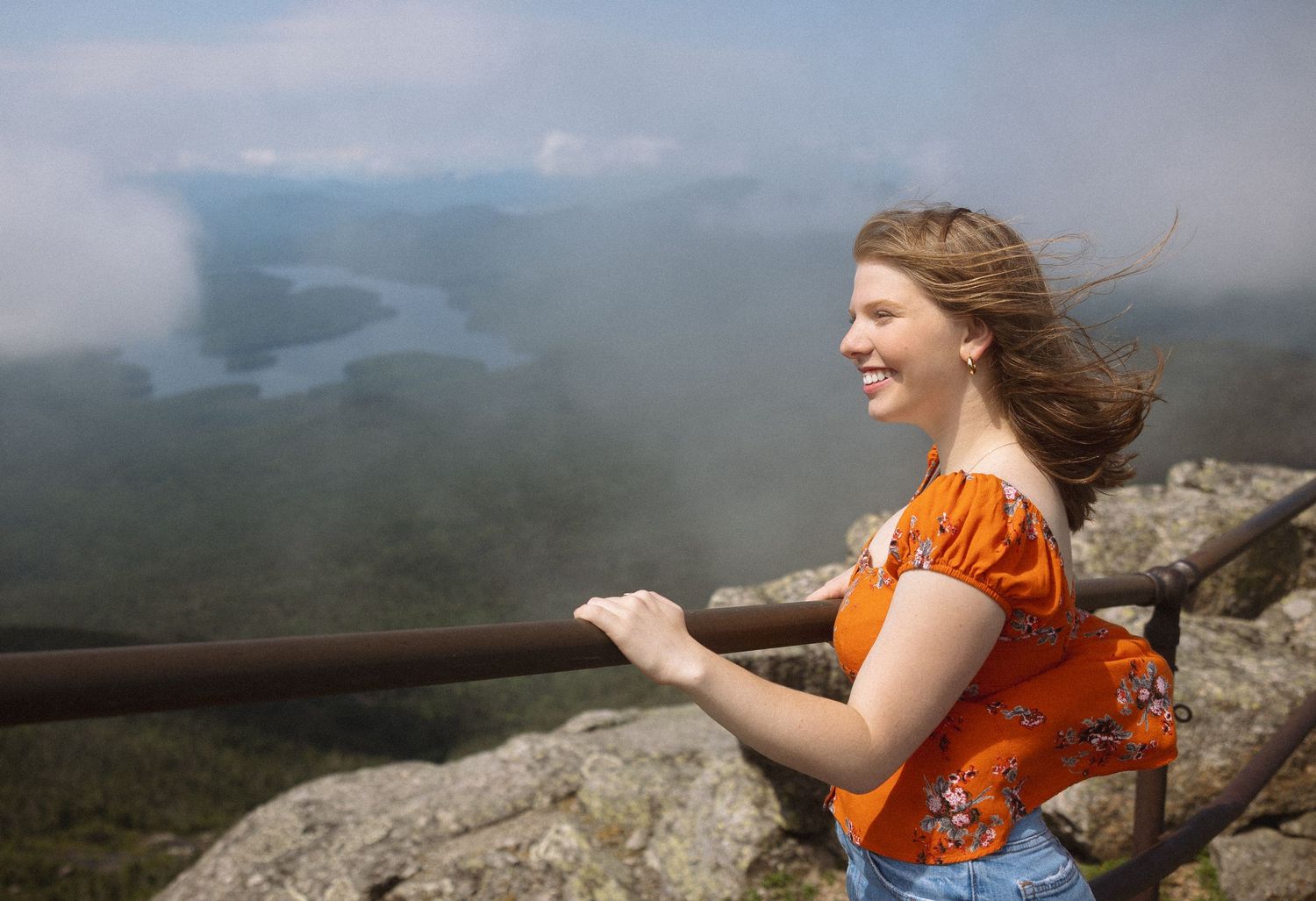 A person in an orange shirt stands on a rocky mountain overlook with windswept hair enjoying the scenic vista below.