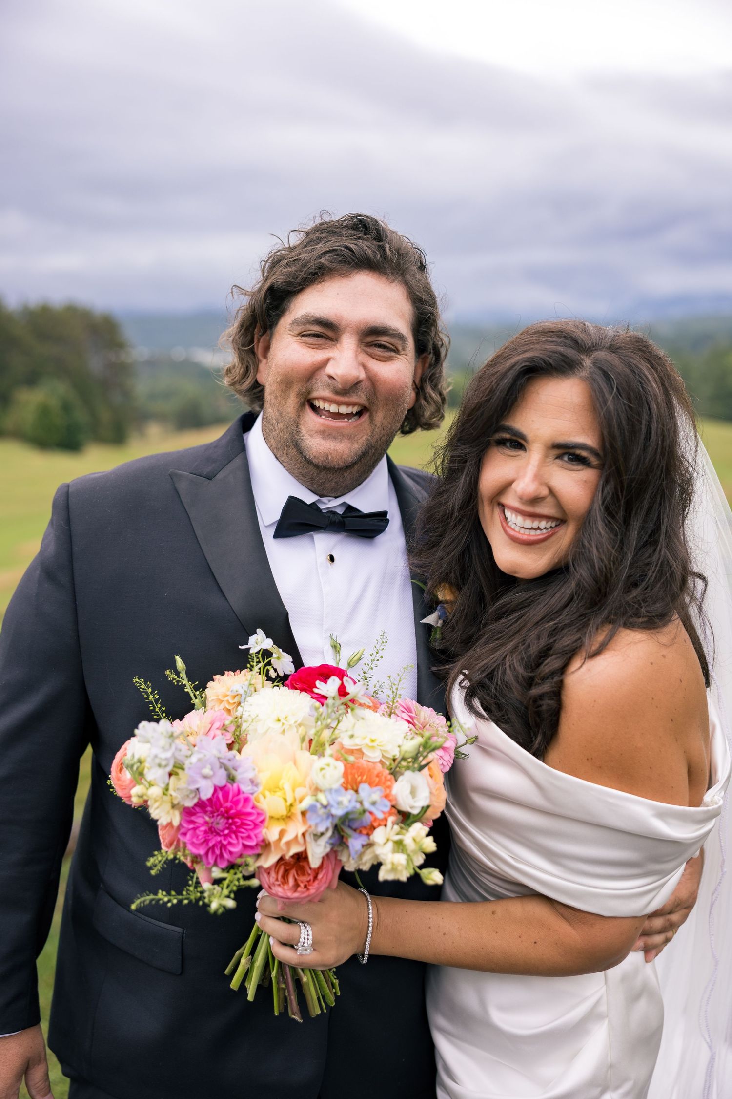 Smiling bride and groom pose outdoors on wedding day, bride holding colorful bouquet, groom in black tuxedo with bow tie.