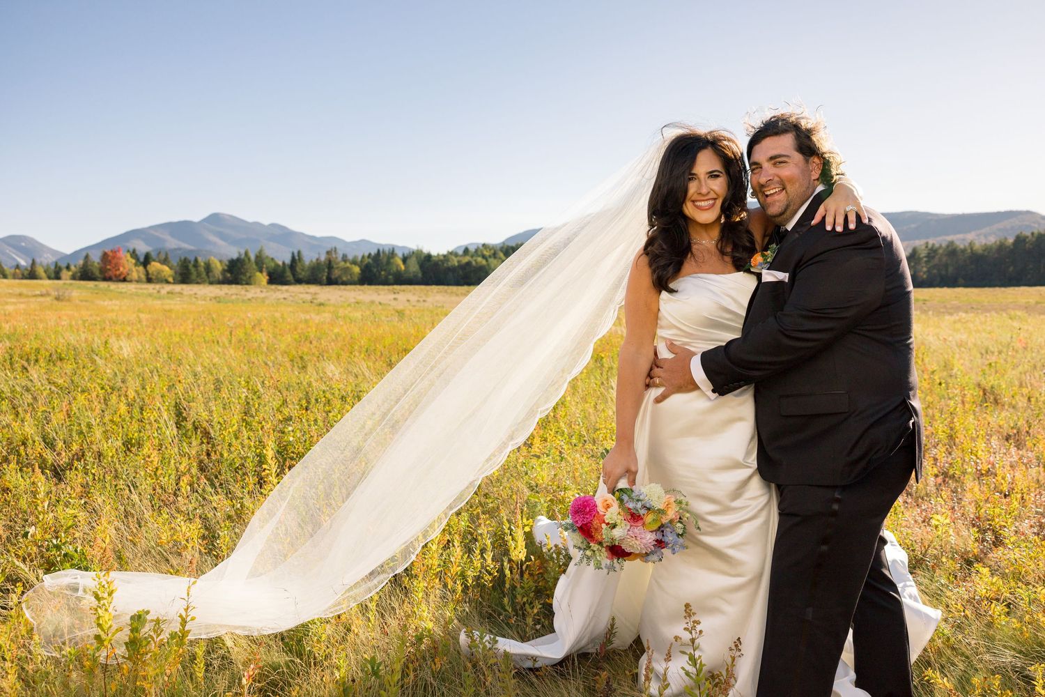 Bride and groom posing in a sunlit meadow with mountains, flowing veil, and colorful bouquet on their wedding day.