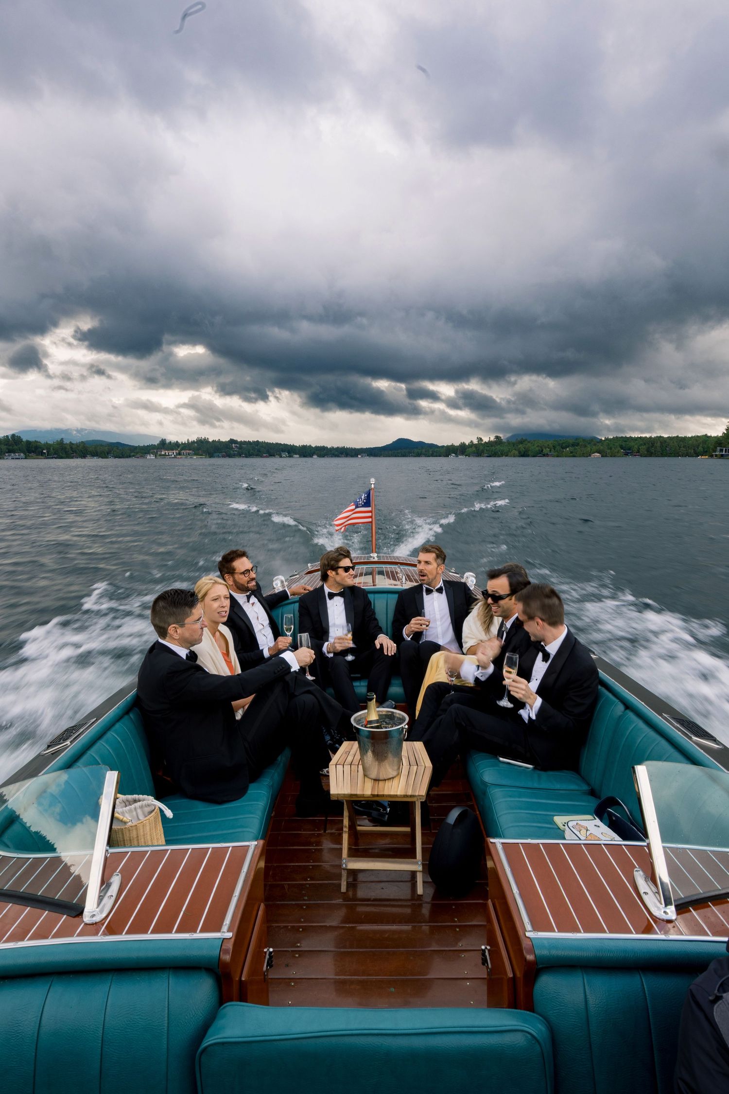 Groomsmen in tuxedos celebrate on a classic wooden speedboat on a lake under dramatic cloudy skies.