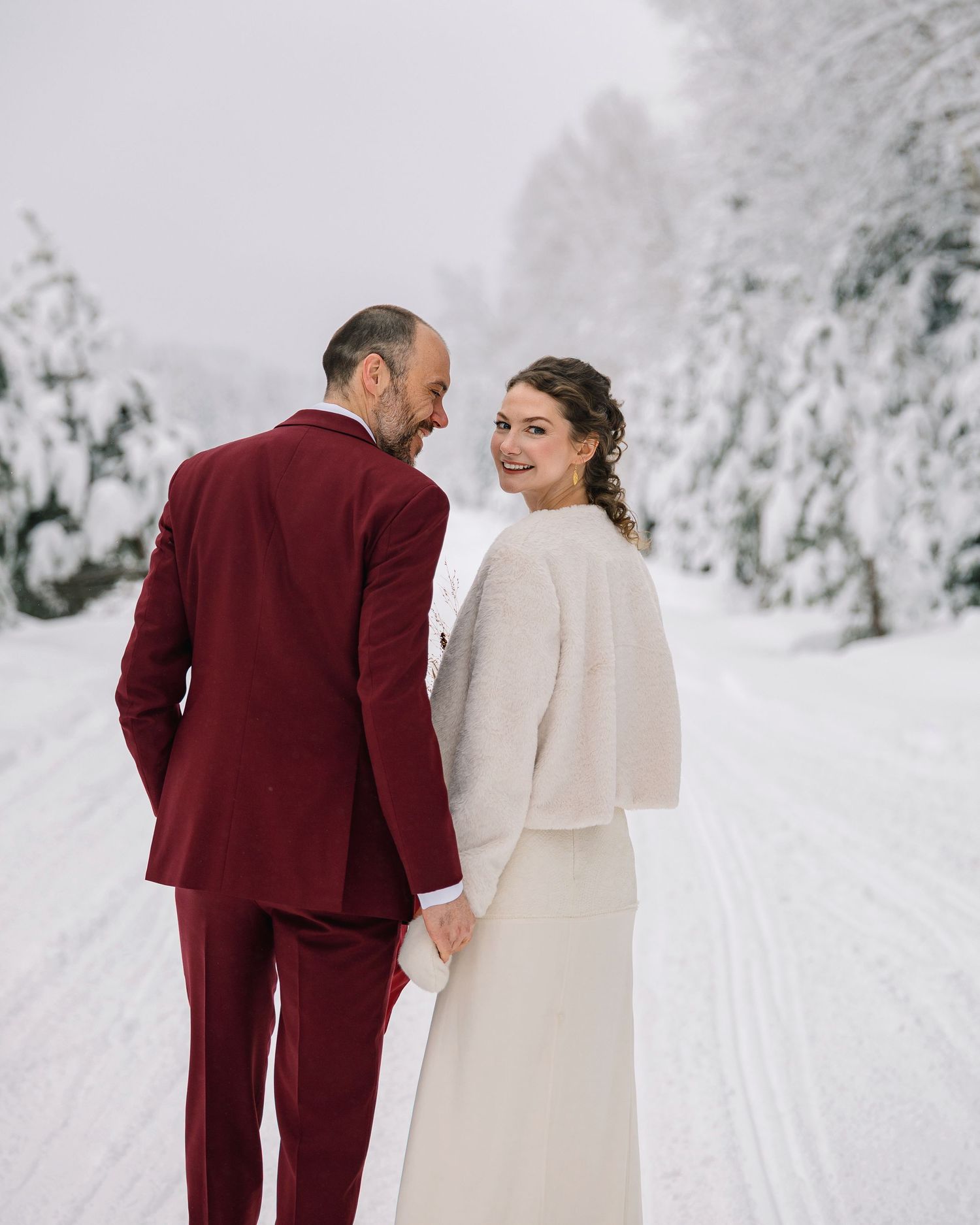A couple in wedding attire stands together in a snowy winter forest landscape with snow-covered evergreen trees.