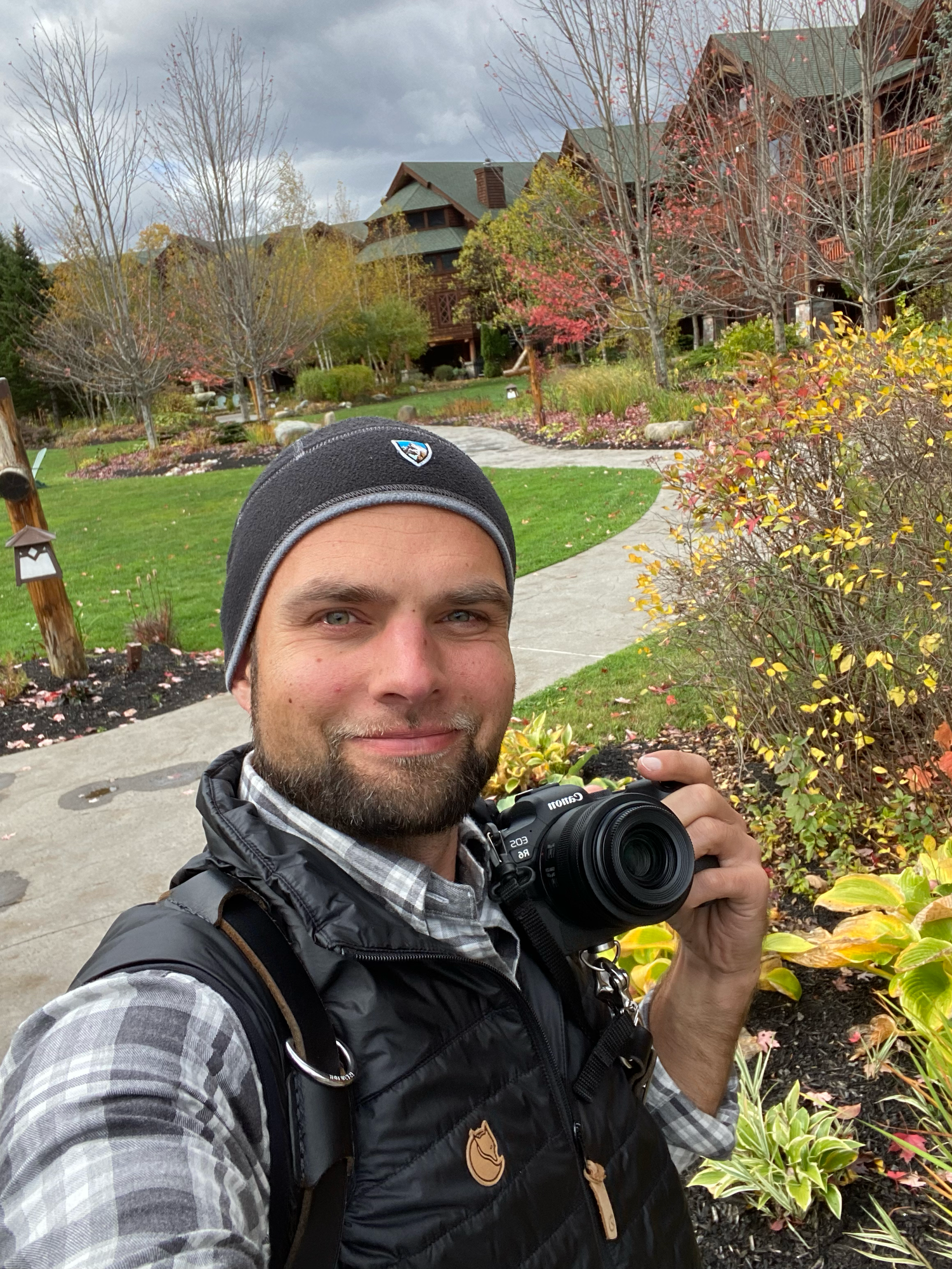 A photographer with camera stands on a winding path through colorful autumn gardens with mountain homes in background.