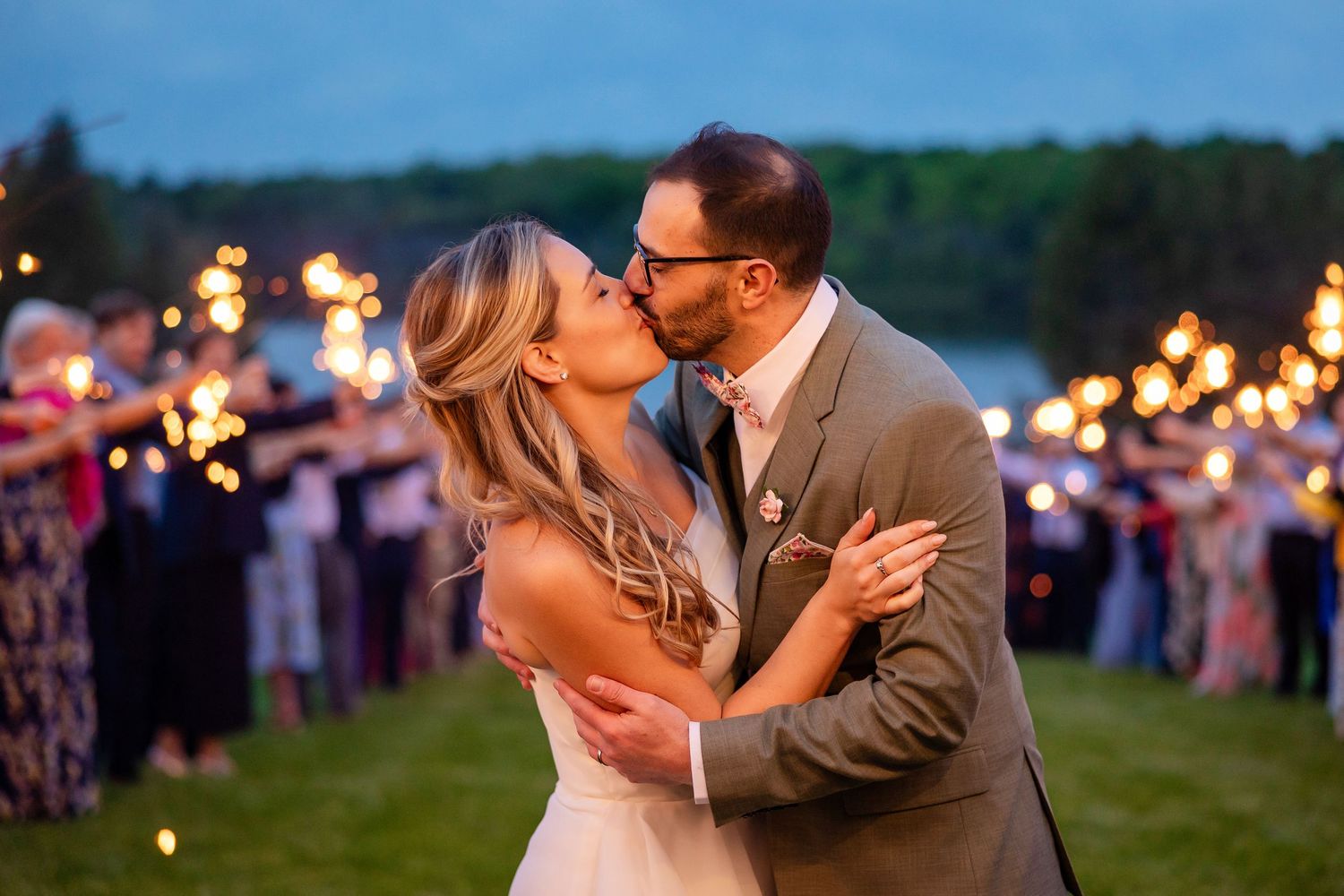 Bride and groom share a romantic kiss surrounded by guests holding sparklers at twilight outdoor wedding reception.