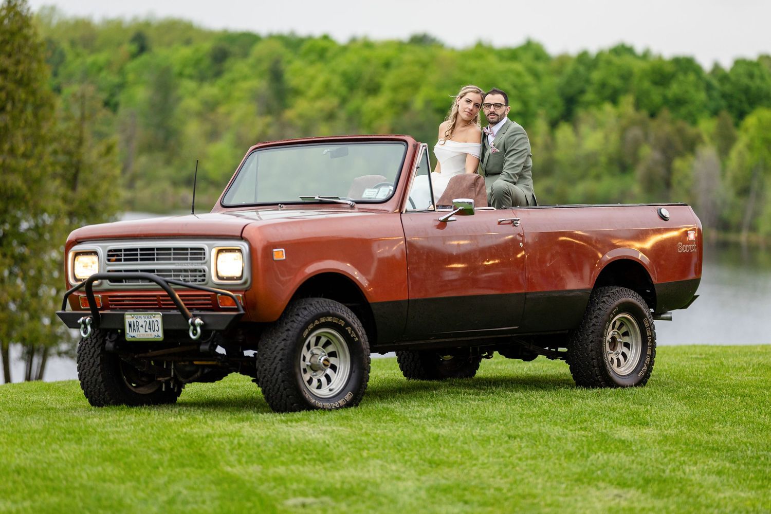 Couple poses in a lifted vintage red International Scout truck on a green lawn near a lake.