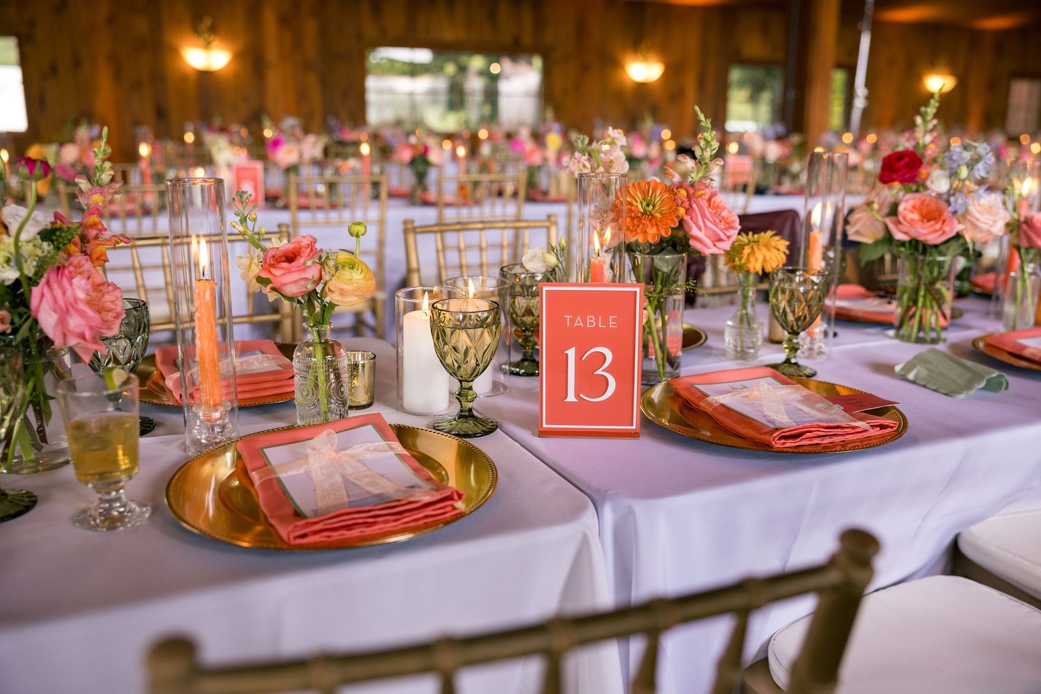 Elegant wedding reception table setting with coral napkins, gold chargers, pink floral centerpieces, and table number 13 sign.