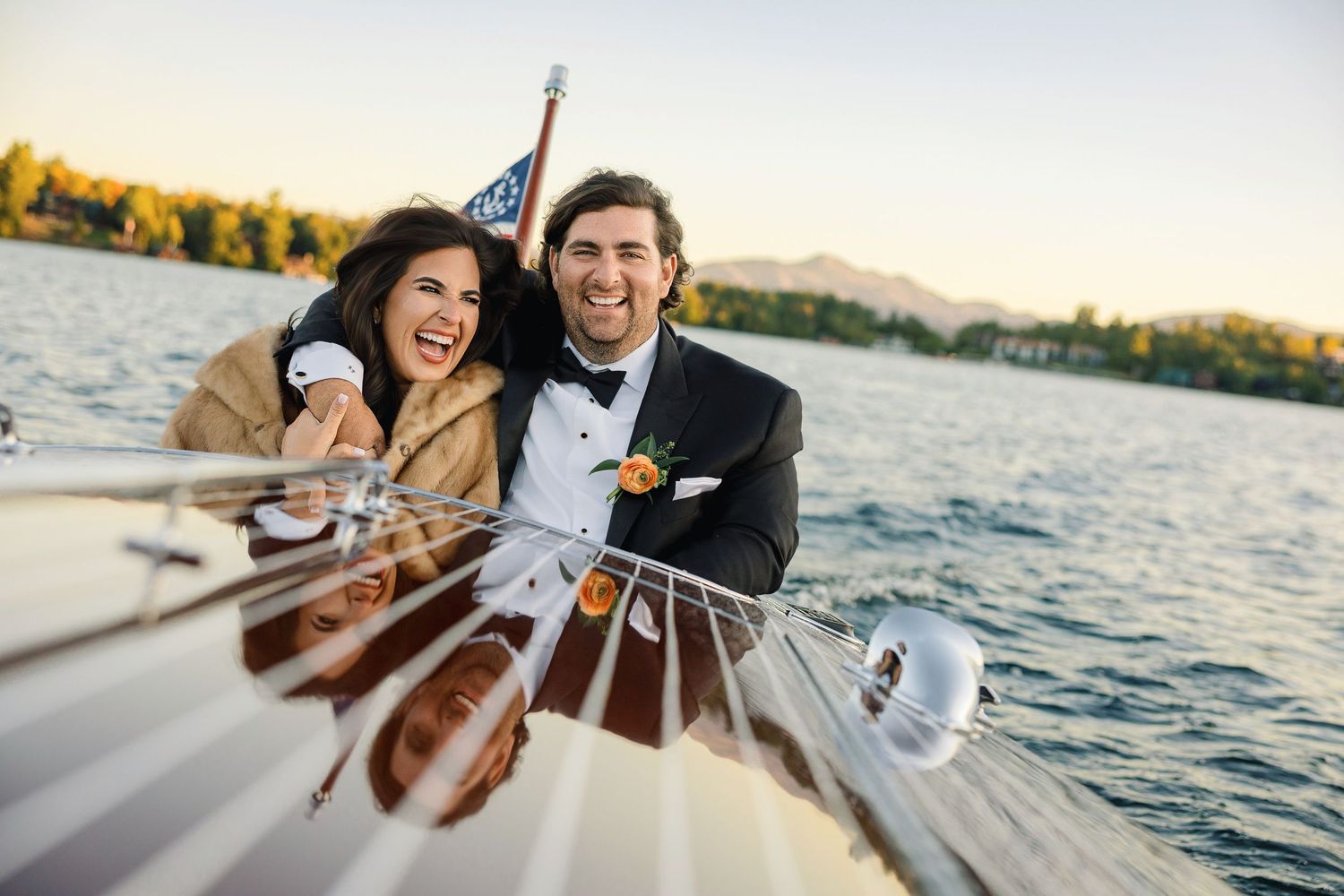 Happy couple in formal wedding attire smiling on a classic wooden boat on a scenic lake at sunset.