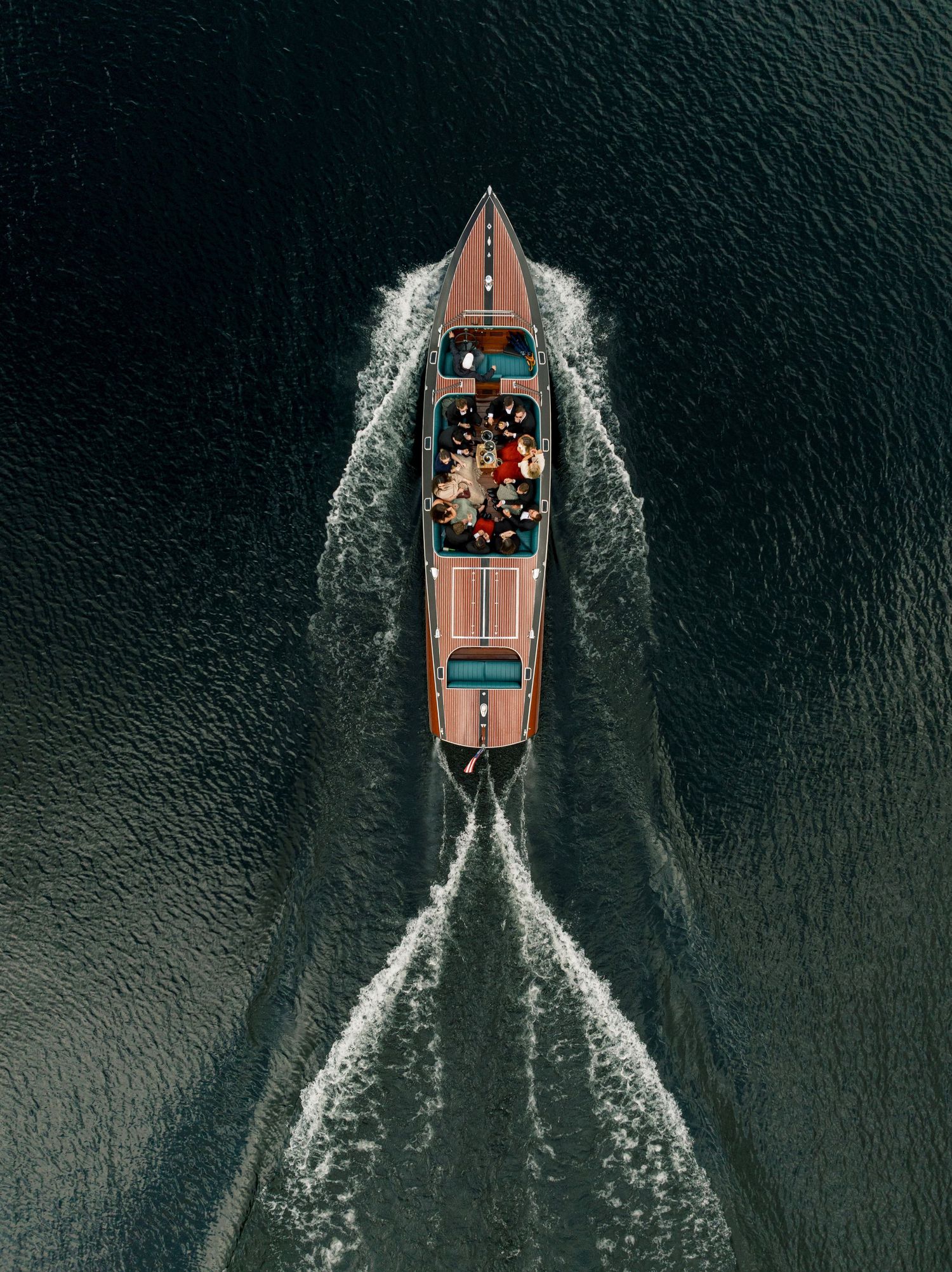 Aerial view of a classic wooden speedboat cutting through dark green water, leaving a white wake trail.
