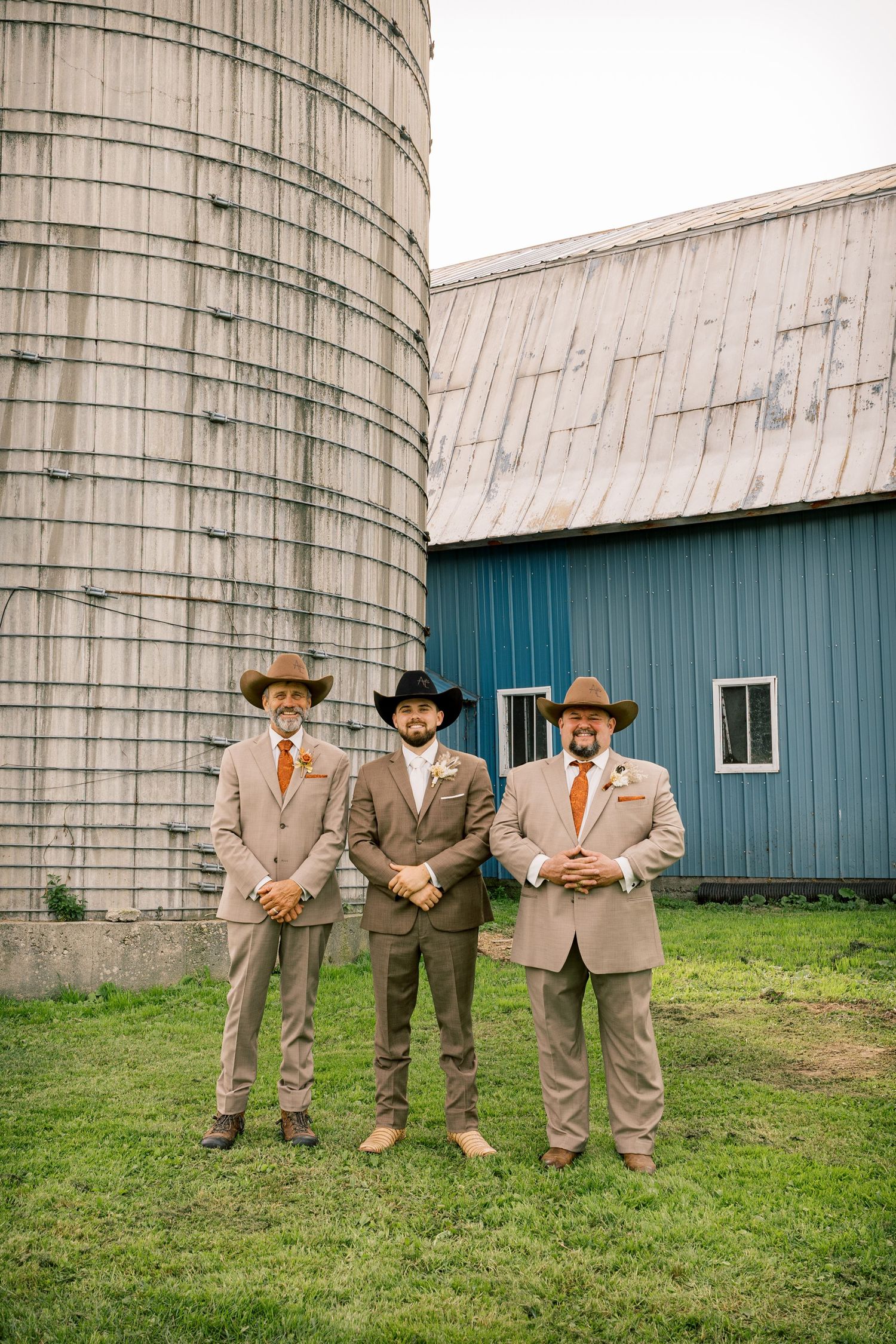 Three groomsmen in tan suits, orange ties and cowboy hats pose on green grass in front of a rustic silo and blue barn.