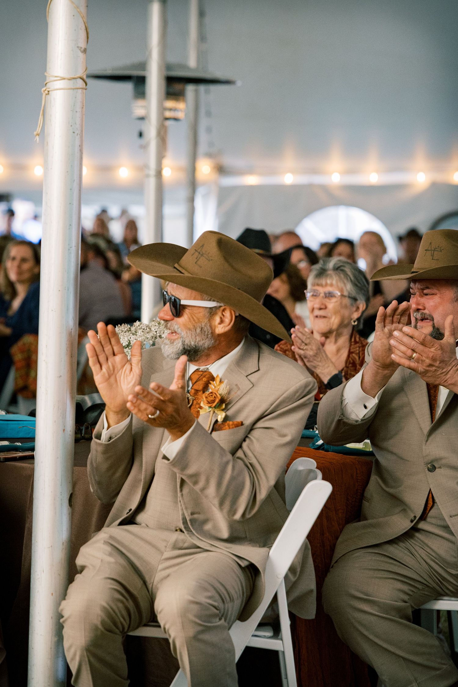 Wedding guests in tan suits and brown hats applaud and smile while seated under a tent with string lights.