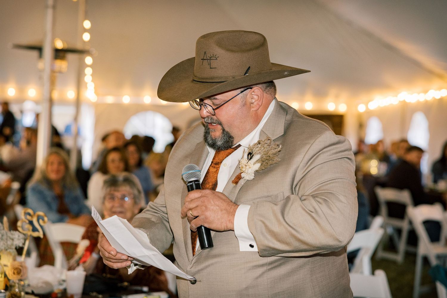 Man in a tan suit and cowboy hat speaks into a microphone while reading notes to seated guests at a tented wedding reception.