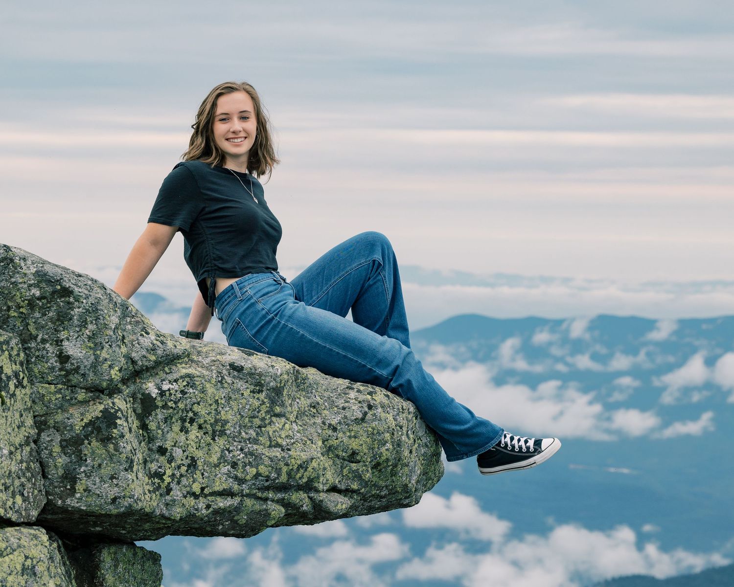 Person in blue jeans sits on rocky mountain ledge overlooking scenic misty valley landscape at sunset.