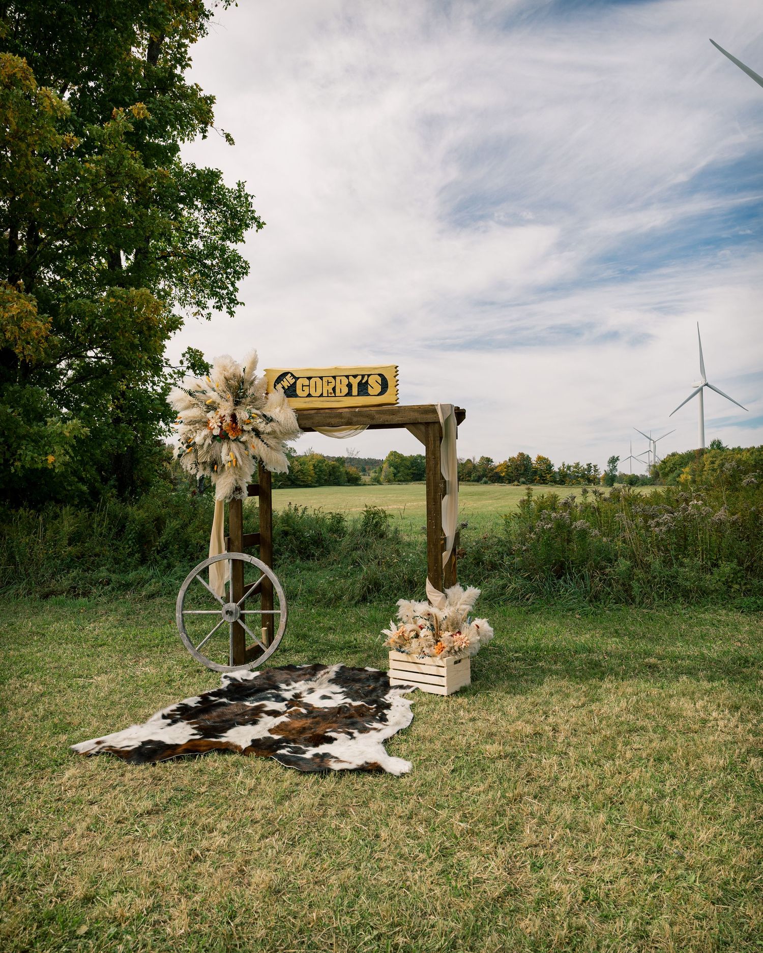 Rustic wooden welcome arch with a Gordy's sign, pampas grass florals, wagon wheel and cowhide rug in a field with wind turbines.