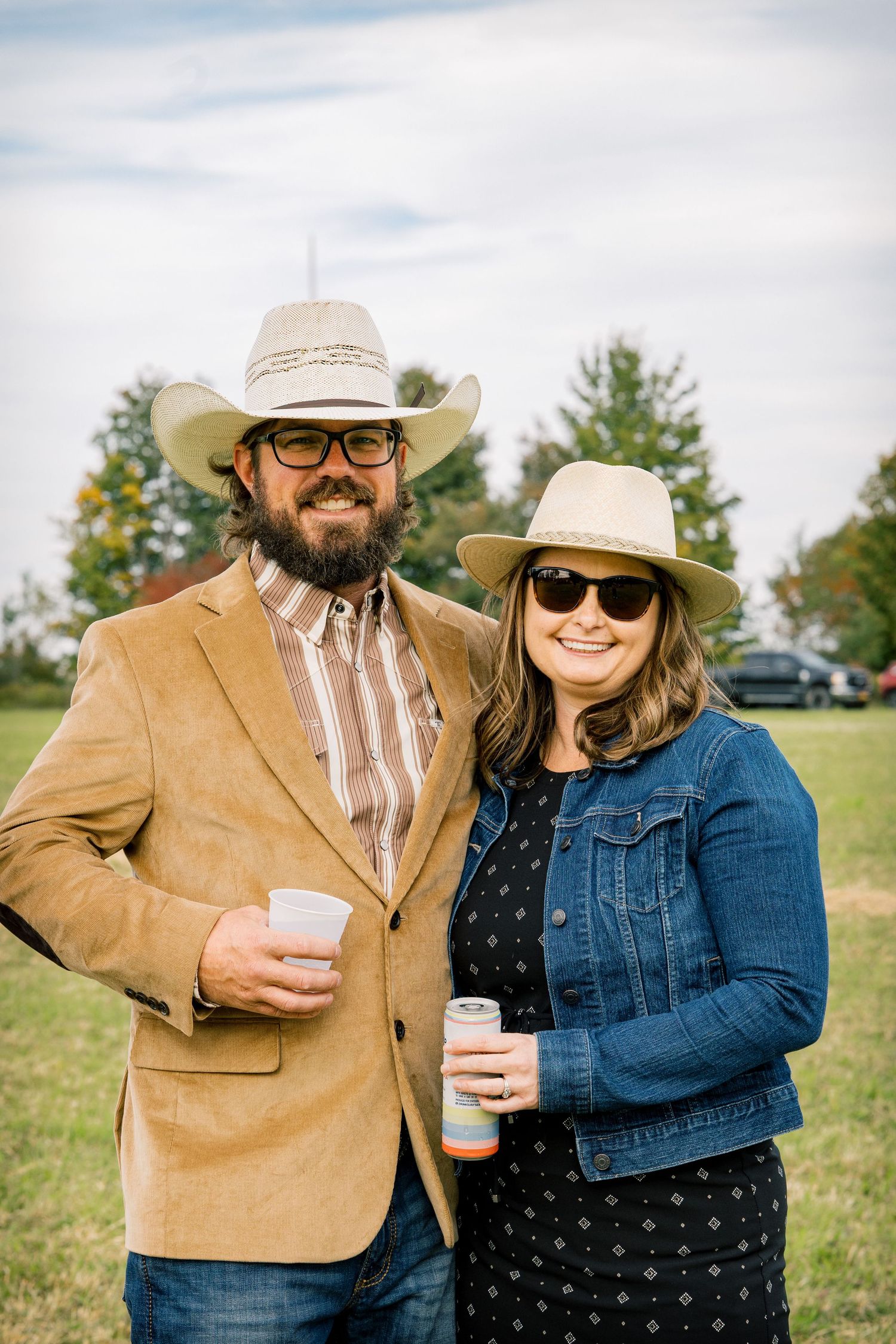 Bearded man in a tan blazer and cowboy hat and smiling woman in a denim jacket and sunglasses hold drinks outdoors at a country gathering.