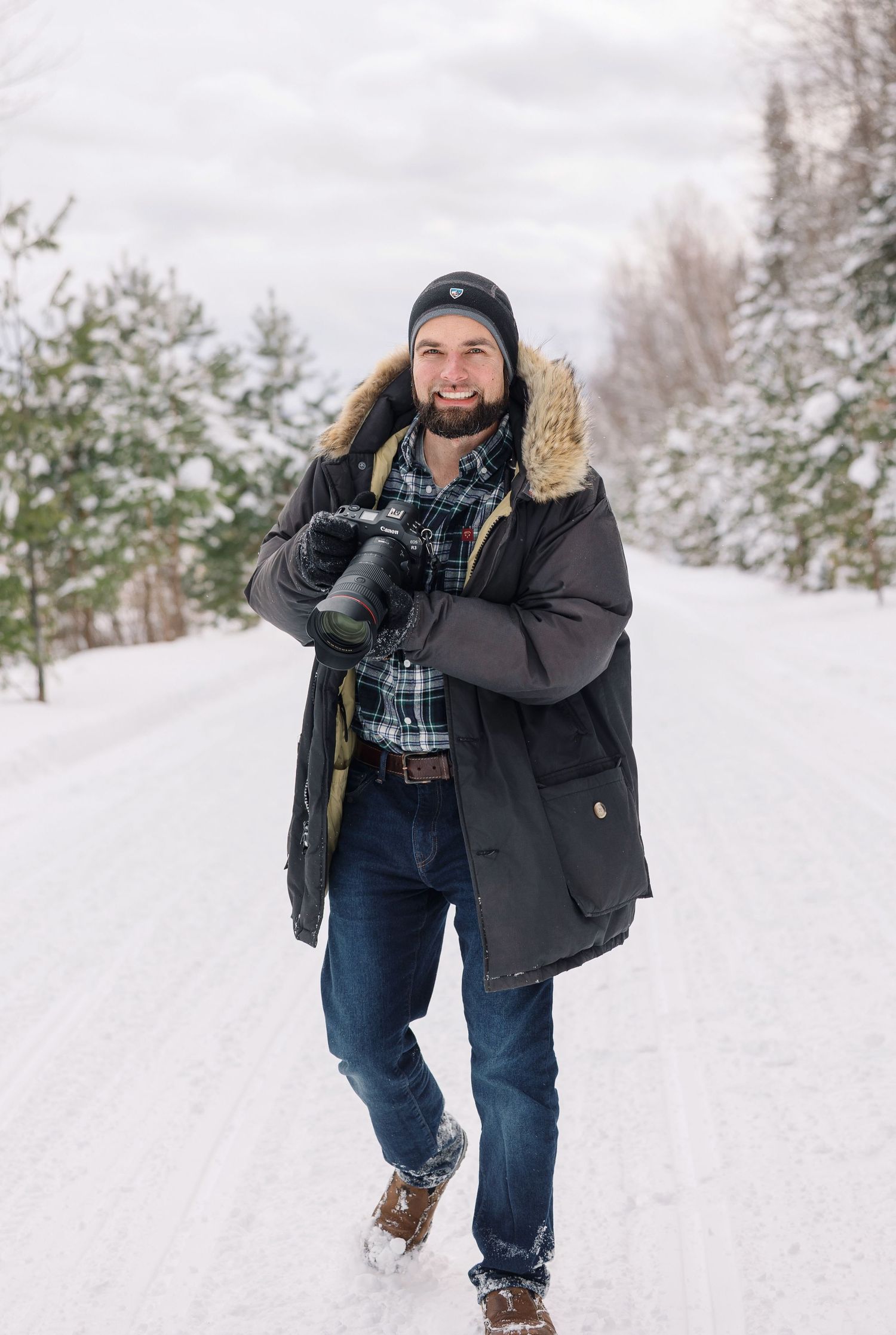 Person in winter attire walking through snowy landscape with evergreen trees in background during winter season.