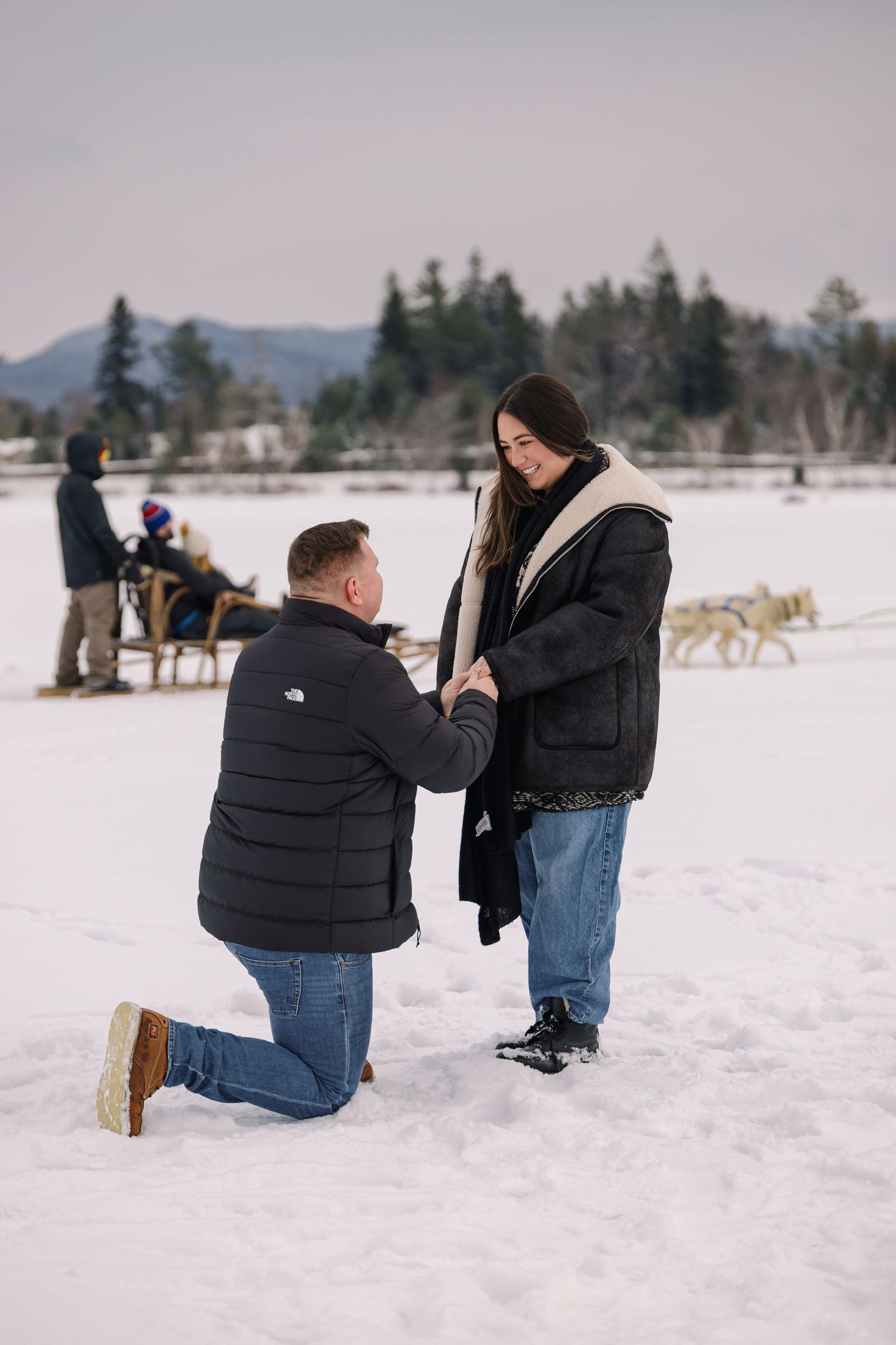 A winter proposal scene on a snowy field with mountains and trees in the distance.