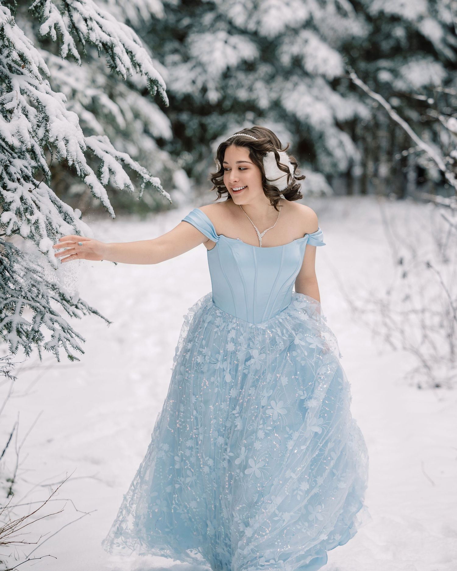 A person in a light blue ballgown poses gracefully in a snowy winter forest setting with snow-covered branches.