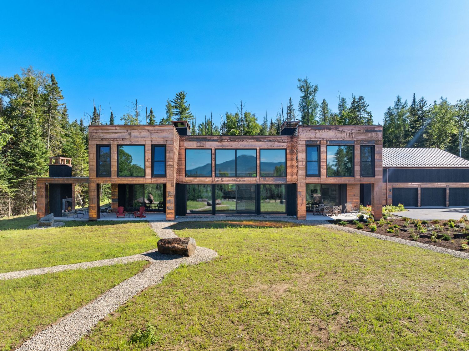 Modern luxury home with glass walls and wood exterior features a fire pit on manicured lawn surrounded by evergreen trees.
