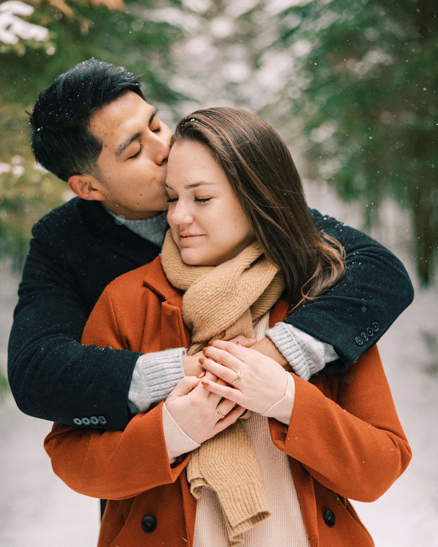 A couple embraces in winter wearing cozy sweaters and coats against a snowy outdoor background.