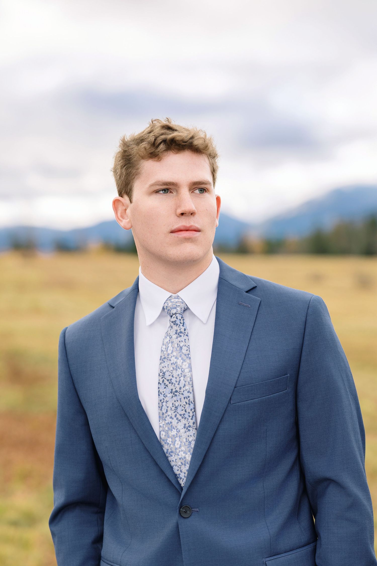 A person in a navy blue suit and floral tie stands in front of a mountain landscape on an overcast day.