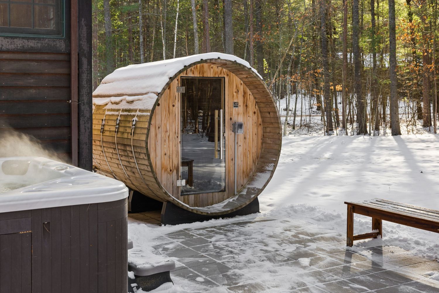 A rustic wooden sauna barrel sits in a snowy winter forest setting with sunlight filtering through the trees.