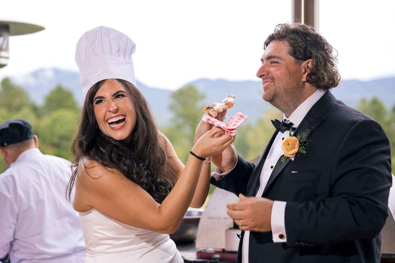 Bride in chef hat and groom in tuxedo laughing while sharing food at outdoor wedding reception.