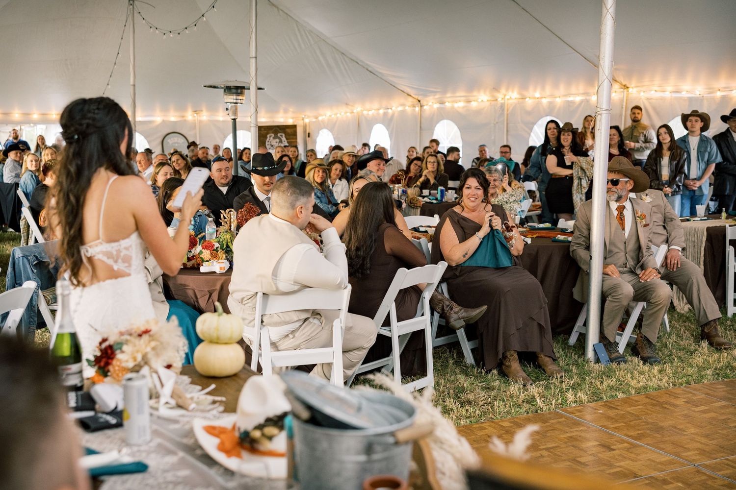 Wedding guests seated at tables under a large white tent listen as a woman in a white dress speaks during a rustic outdoor reception.