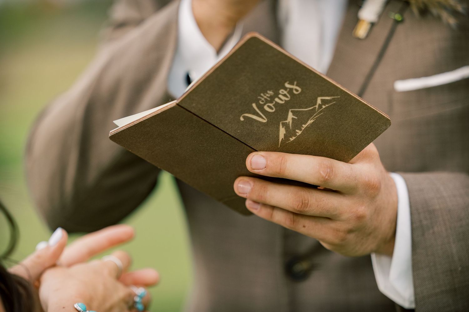 A man in a tweed suit reads from a brown vows booklet while holding hands with his partner during a wedding ceremony.