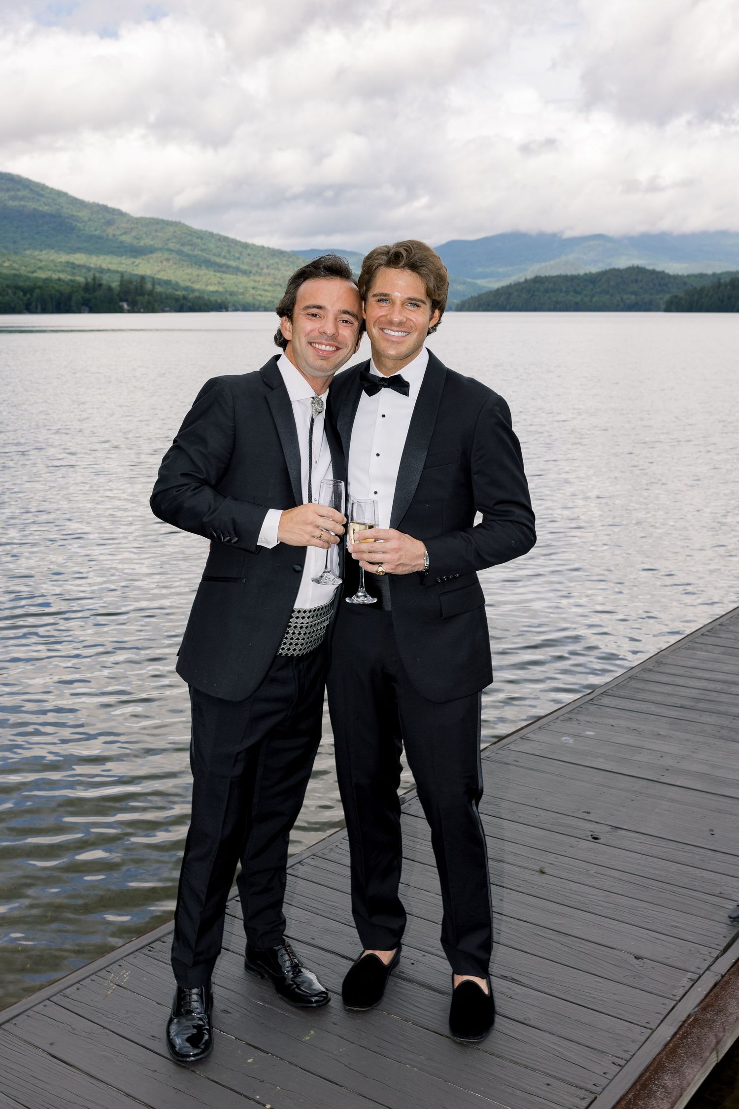 Two men in tuxedos holding champagne glasses on a dock with a scenic lake and mountains backdrop.
