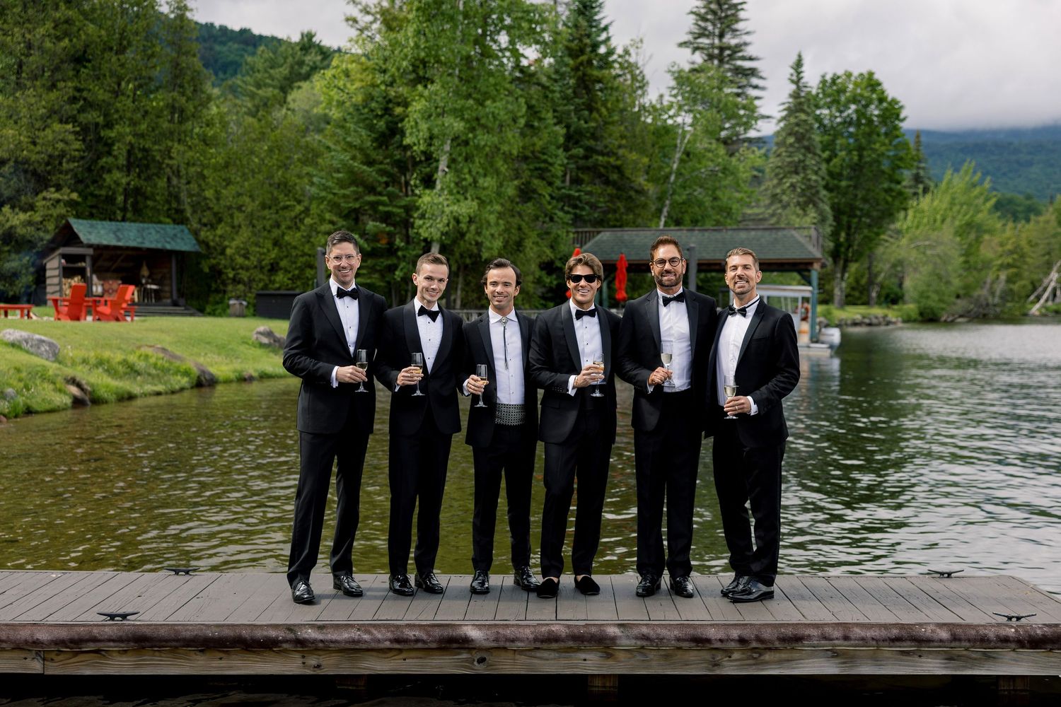 Six groomsmen in black tuxedos pose on a lakeside dock surrounded by lush green mountains at a wedding.