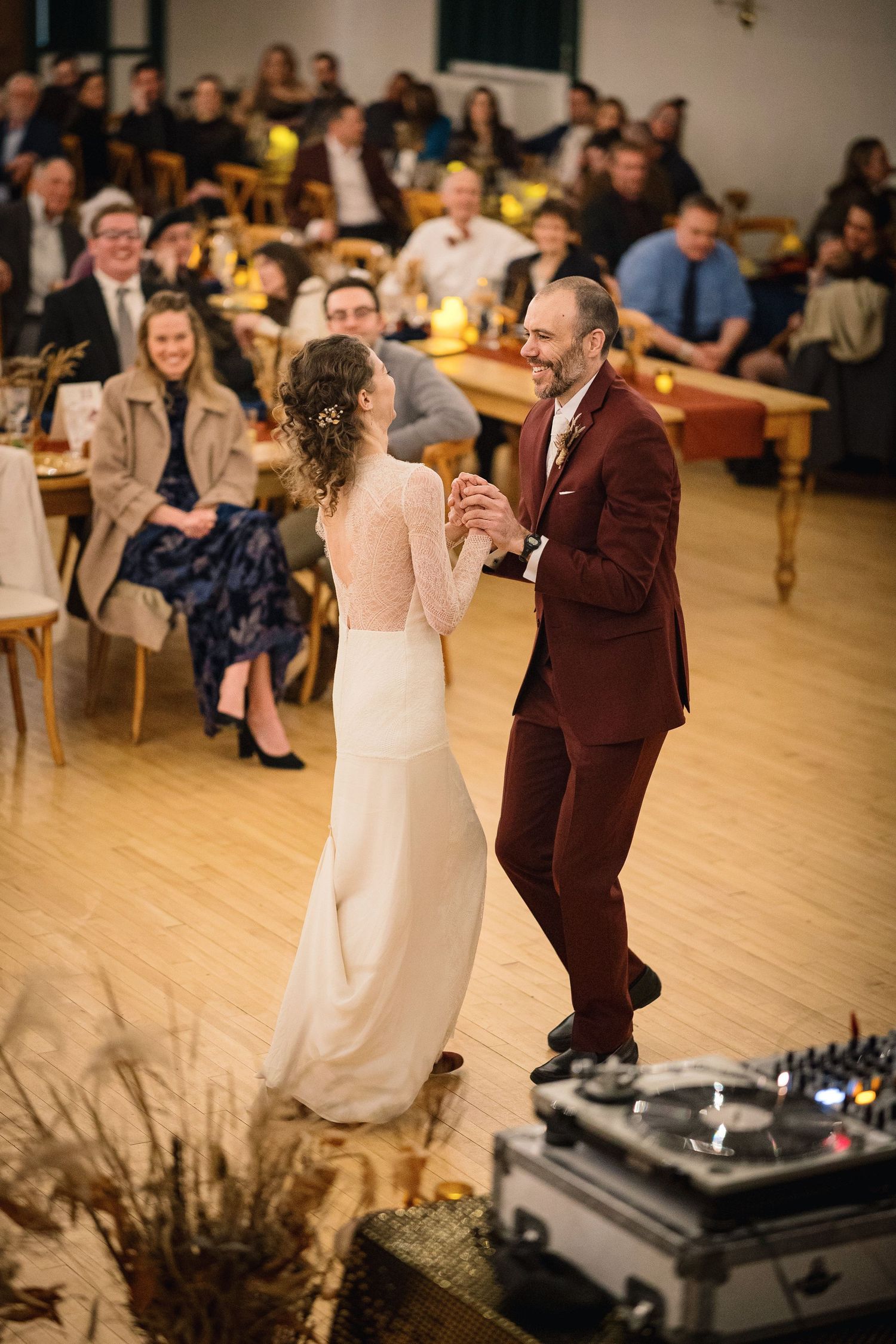 A couple shares their first dance at a wedding reception with guests seated at tables and a DJ setup in the foreground.