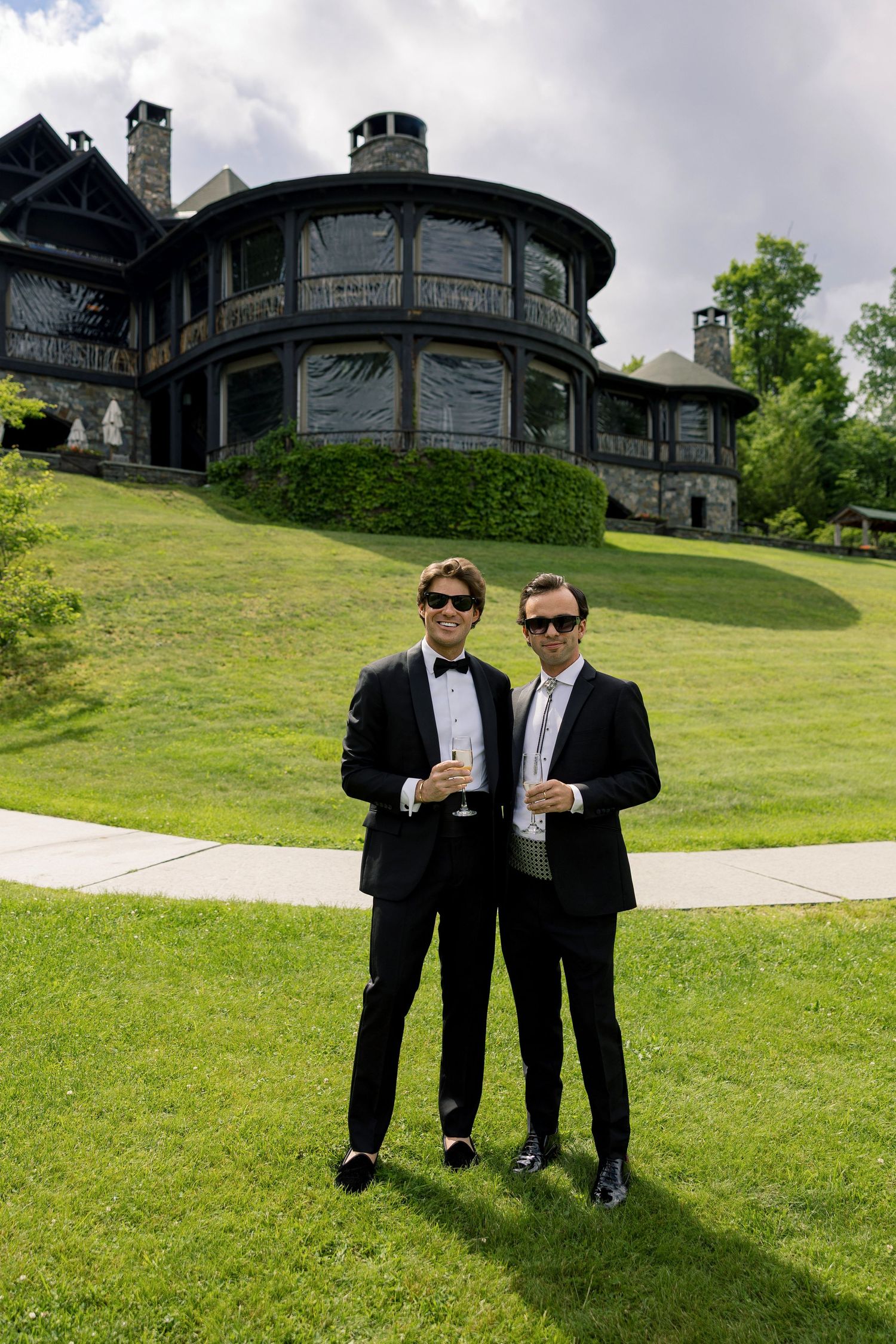Two men in black tuxedos holding champagne glasses stand on green lawn before a grand estate.