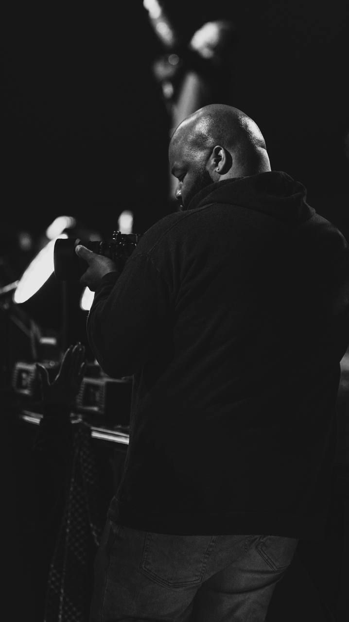 A performer in a dark jacket stands at a microphone on stage, captured in black and white from behind.