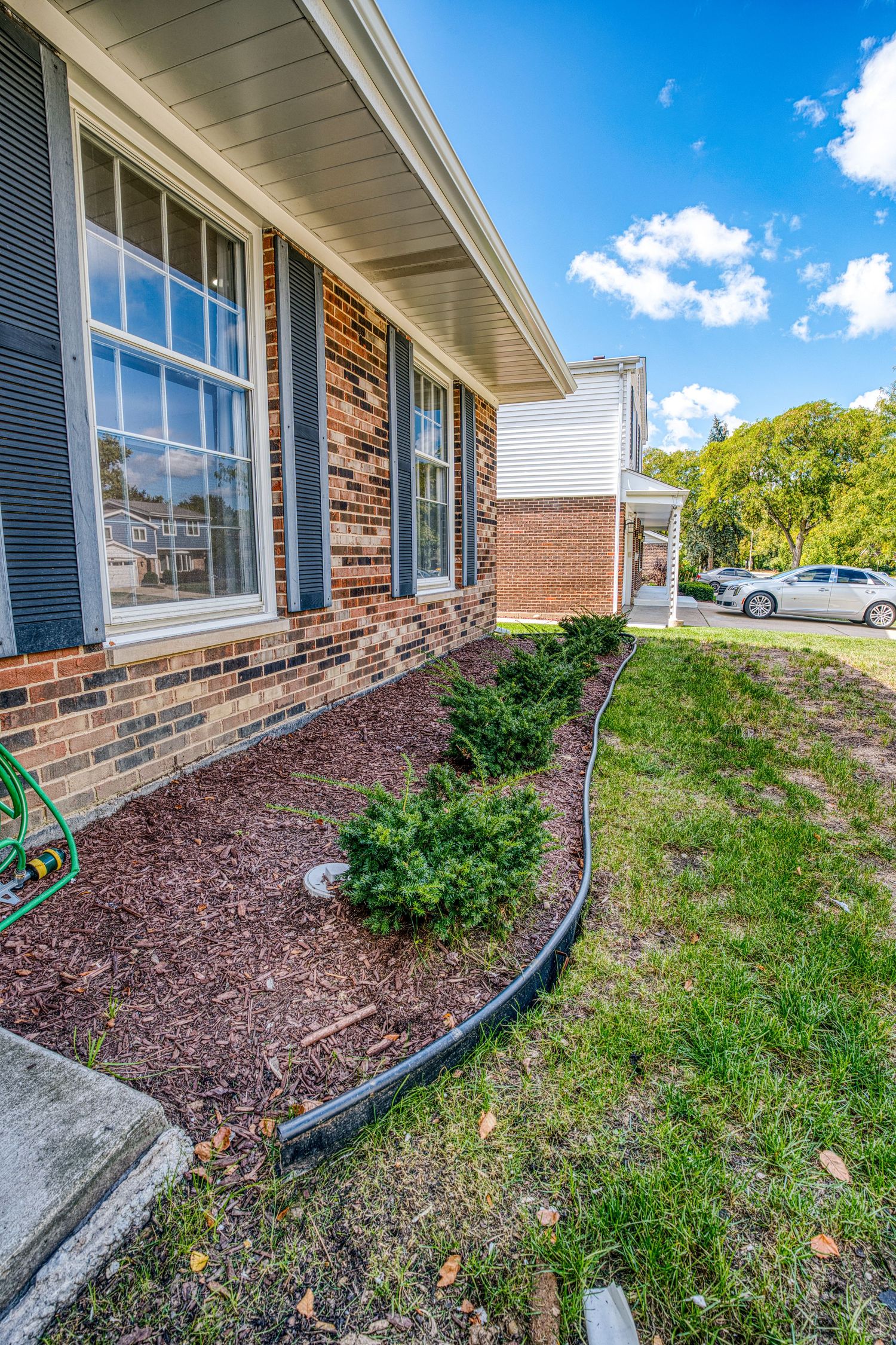Landscaped front garden bed with mulch and shrubs along a brick house exterior with white trim and black shutters.