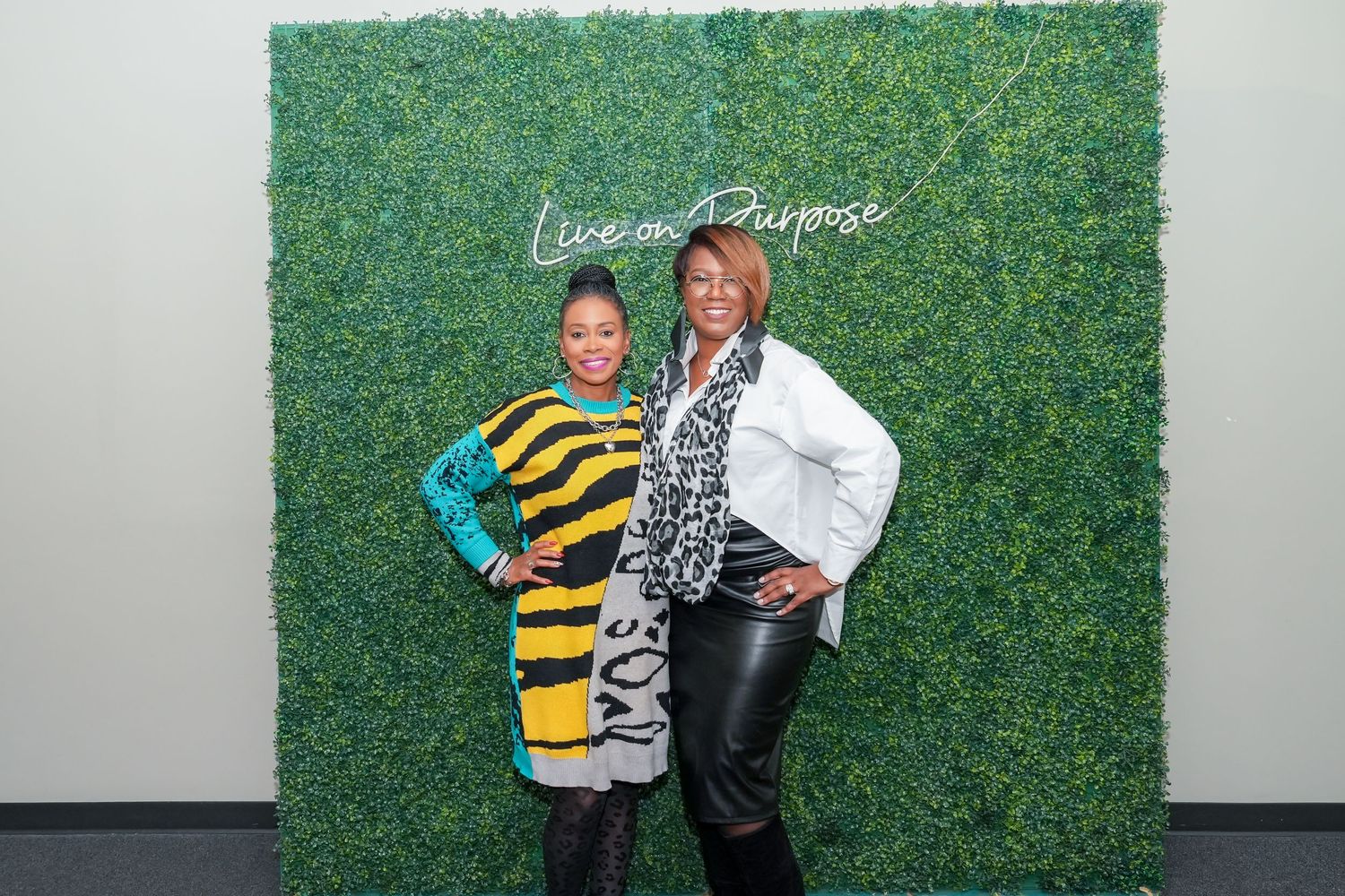 Two people embrace in front of a green hedge wall at a social event.